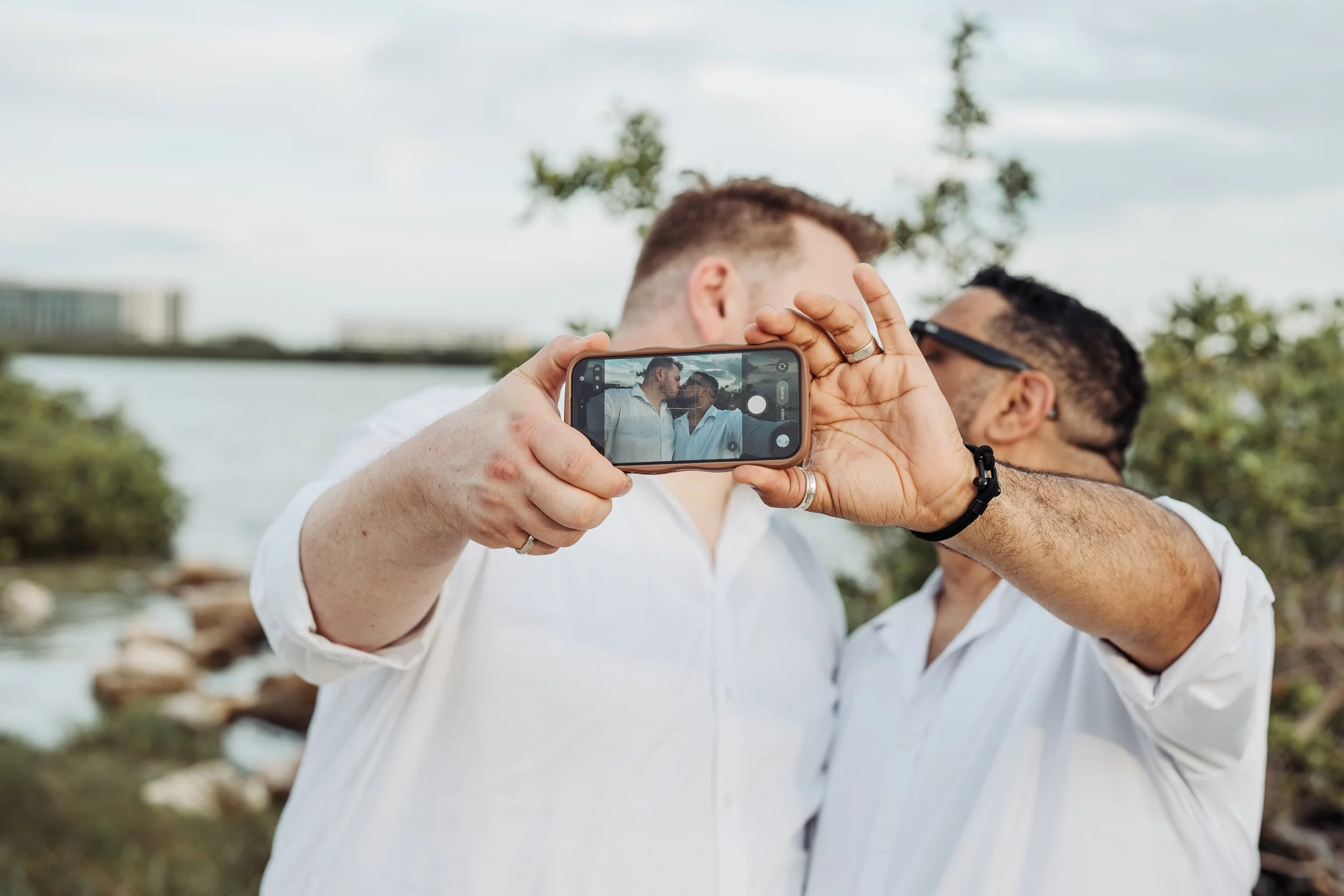 Two men are taking a selfie together outdoors near a body of water, with trees and a bridge in the background. One man is holding a phone, capturing their photo.