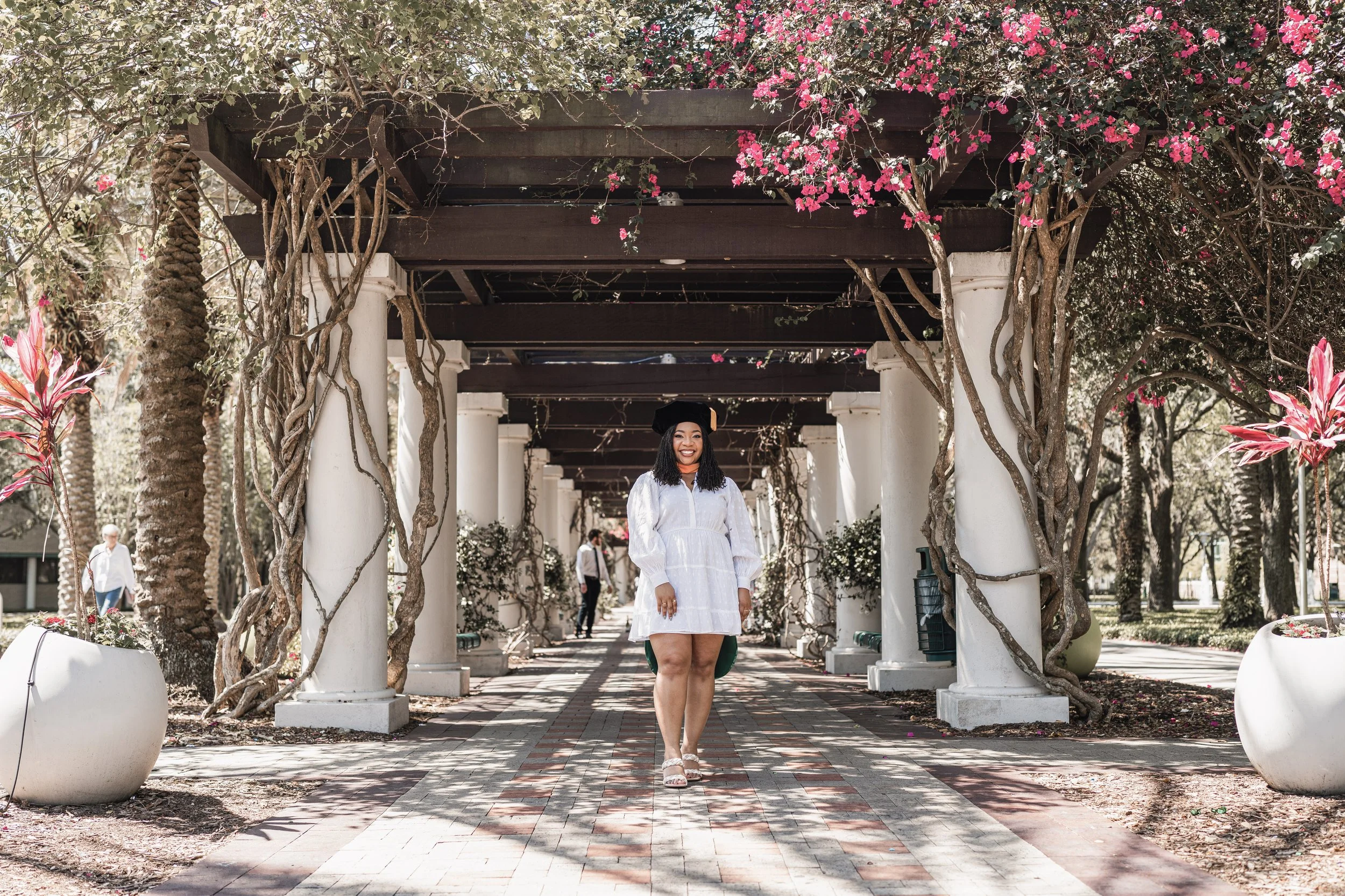 A young woman in a white dress and graduation cap smiling while walking under a pergola with pink flowers and twisting vines, in an outdoor park setting.