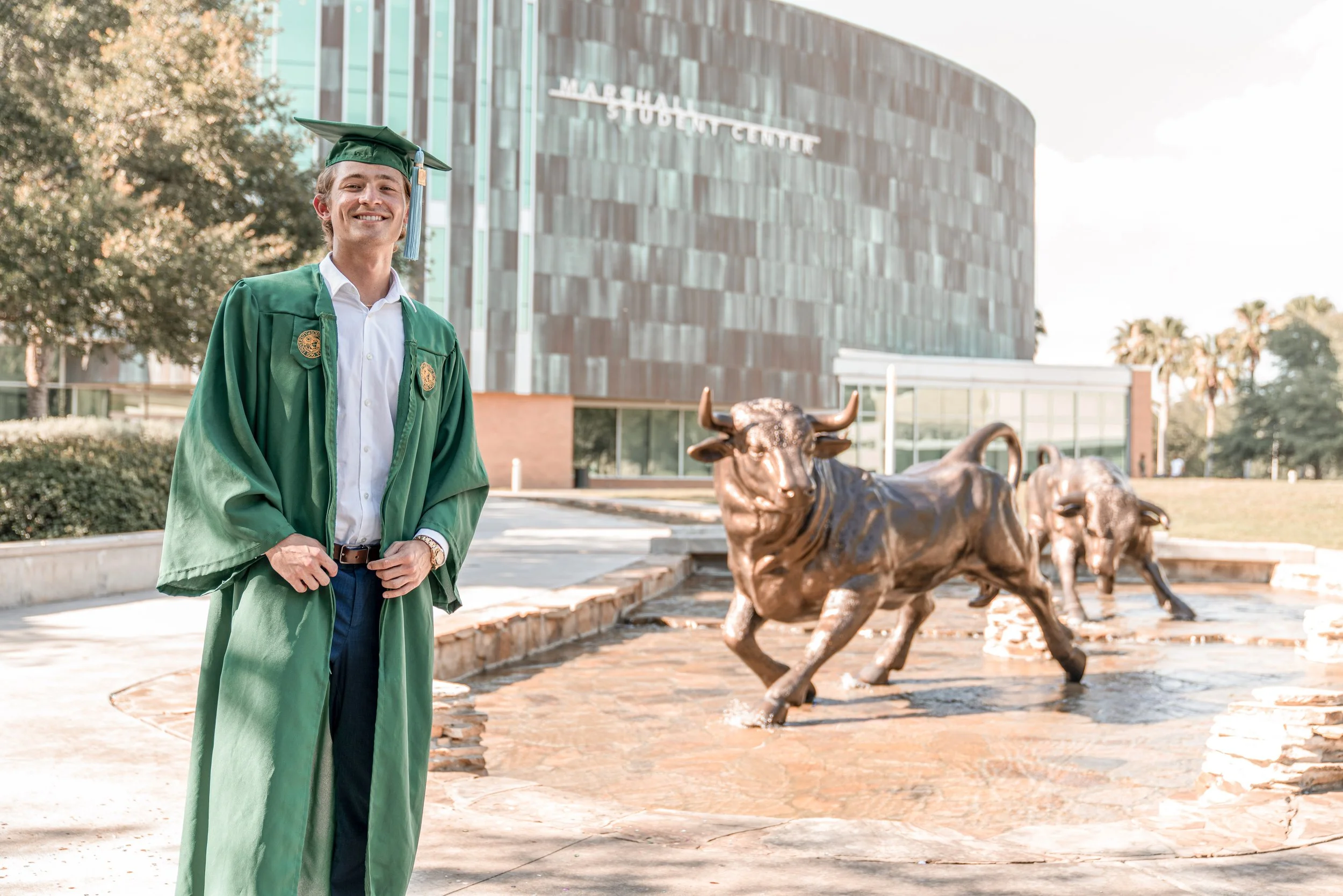 A young man wearing a green graduation gown and cap stands smiling in front of a bronze sculpture of running bulls outside a modern building with glass windows and a sign that reads 'Jade Hub Stadium Center'.
