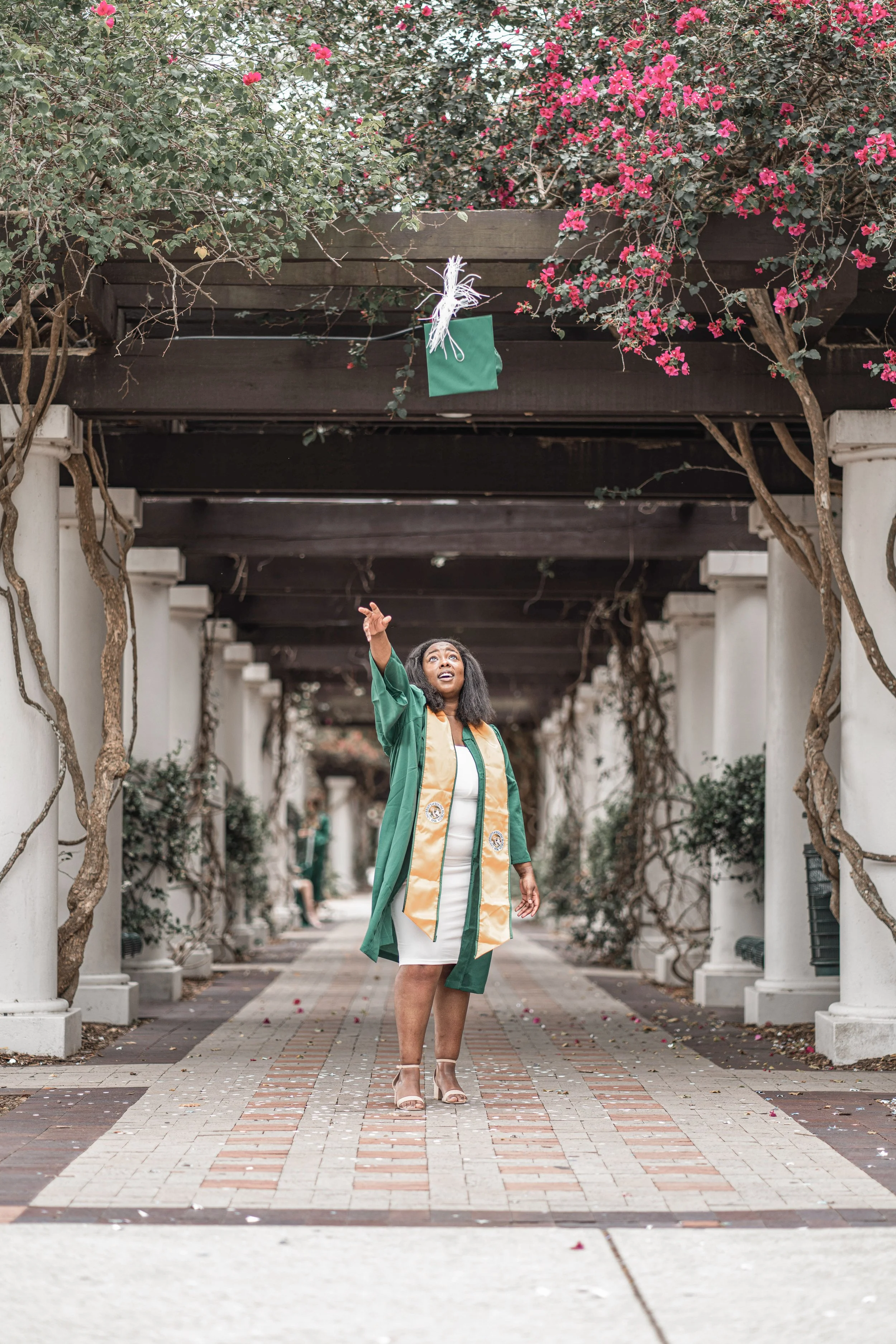 A woman wearing a graduation gown and honor cords is standing on a patterned brick pathway under a pergola with flowering purple and pink bougainvillaea vines, reaching up towards a green gift bag hanging from a wire.