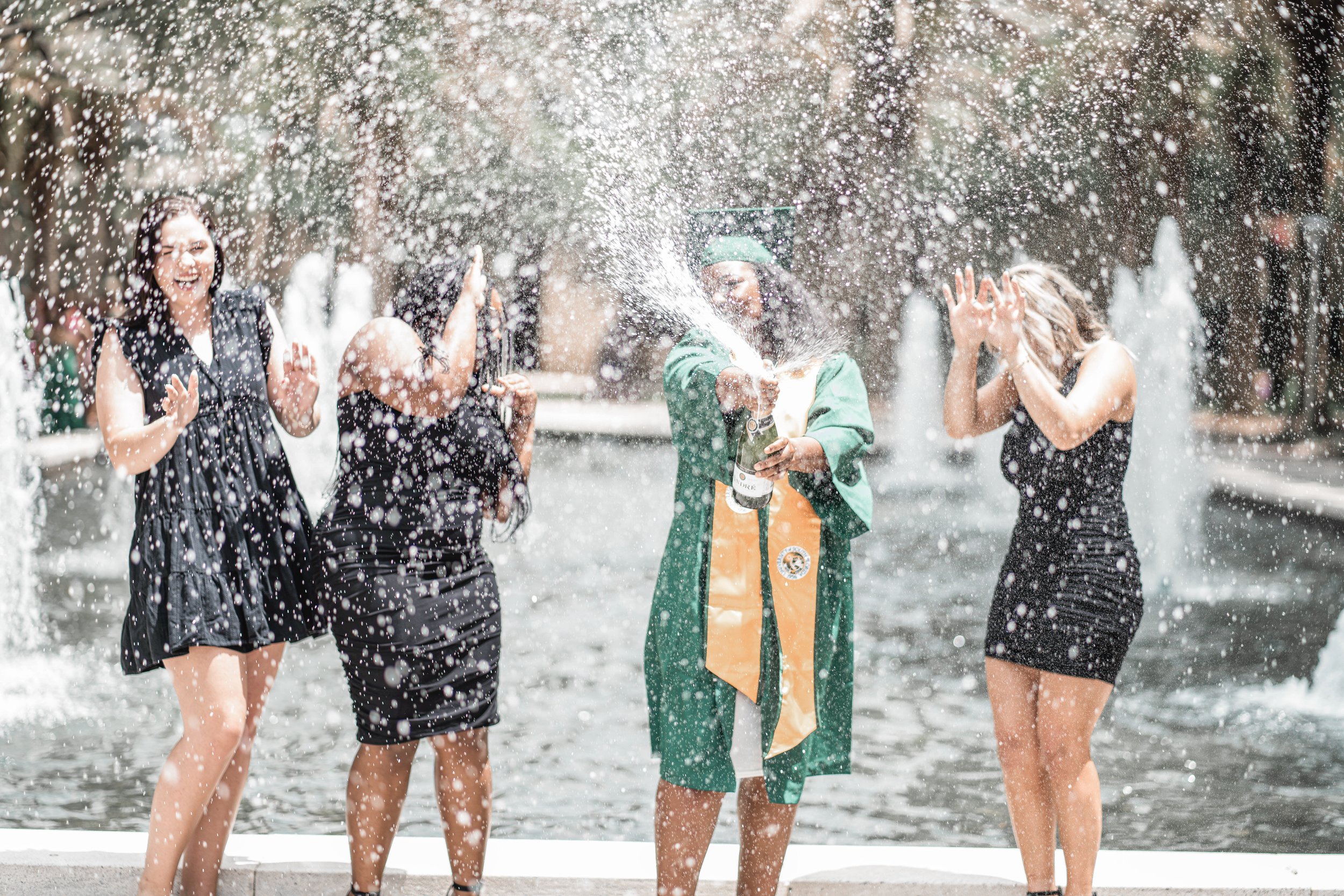 Graduate in cap and gown celebrating with friends at a fountain, spraying water, on a sunny day.