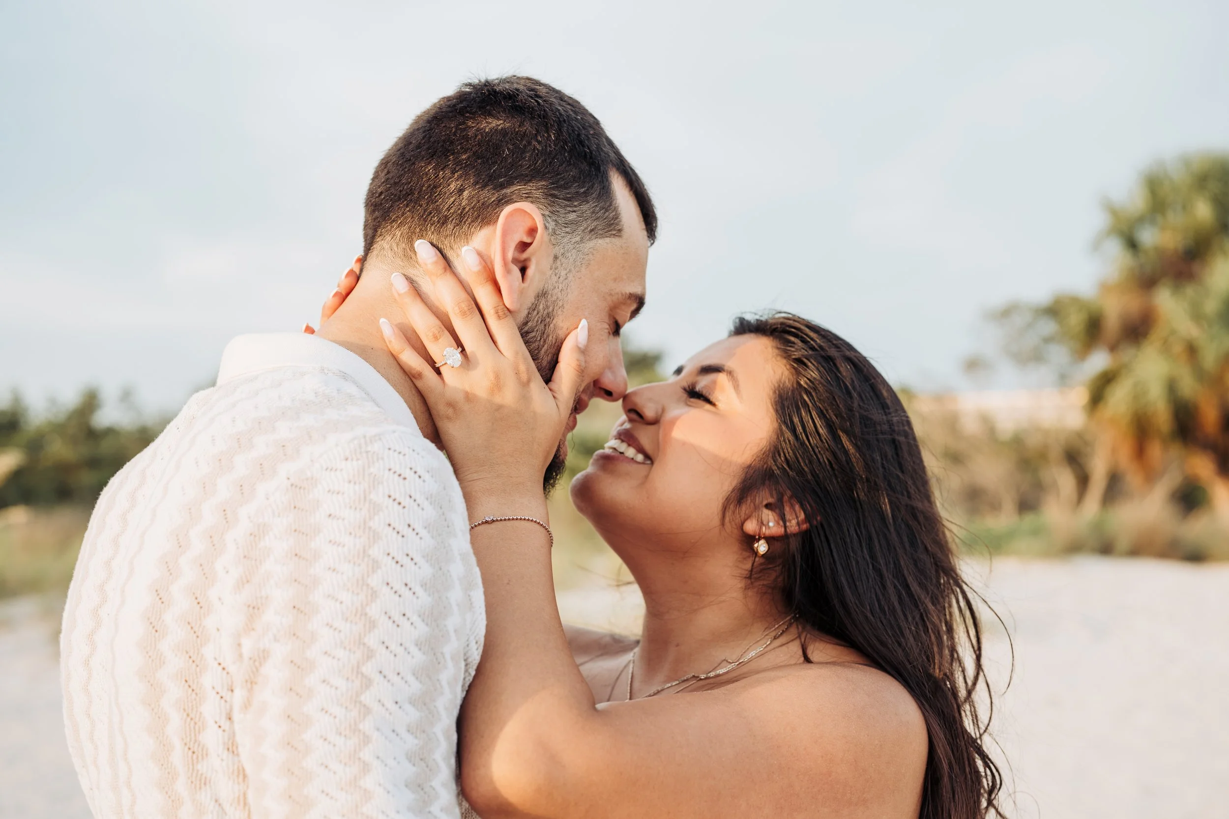 A couple is embracing outdoors, close-up of their faces and upper bodies, with the woman touching the man's face, both smiling with eyes closed, during a sunny day with trees in the background.