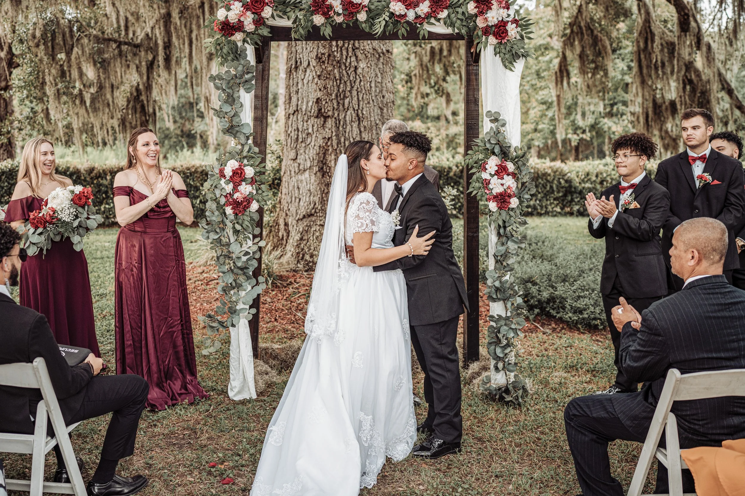 A couple getting married and kissing under a floral wedding arch outdoors during daytime, surrounded by friends and family.