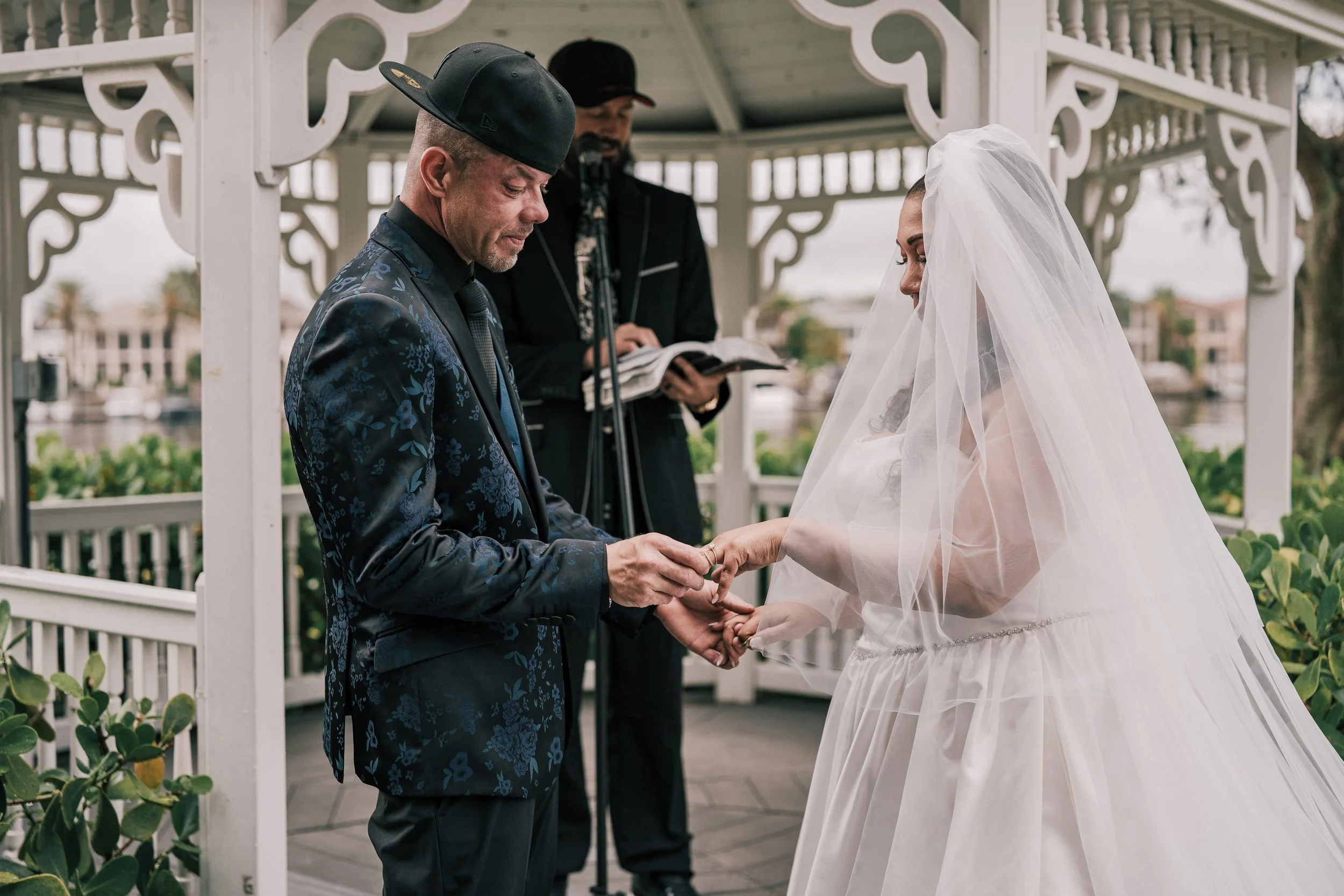 A bride and groom holding hands during their wedding ceremony outdoors under a white gazebo, with an officiant reading, on a cloudy day.