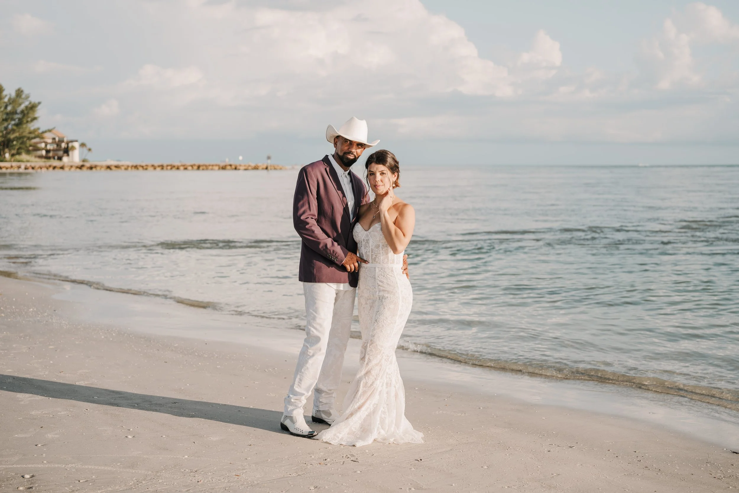 A couple in wedding attire standing on the beach with the ocean in the background, the man wearing a cowboy hat and the woman in a white lace dress.