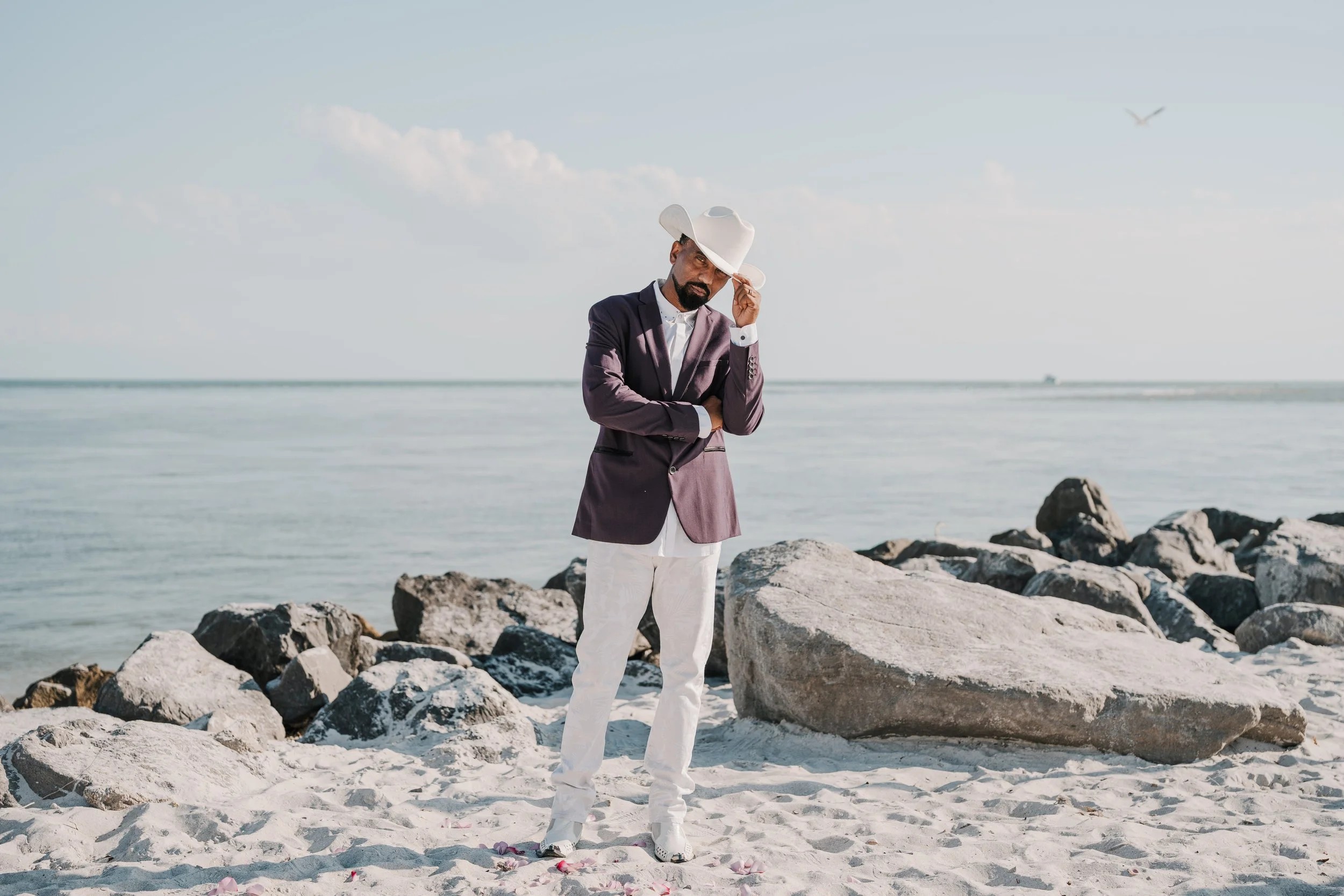 A man in a purple blazer, white shirt, white pants, and white shoes standing on a rocky beach with white sand, during daytime, tipping a white cowboy hat and looking at the camera.