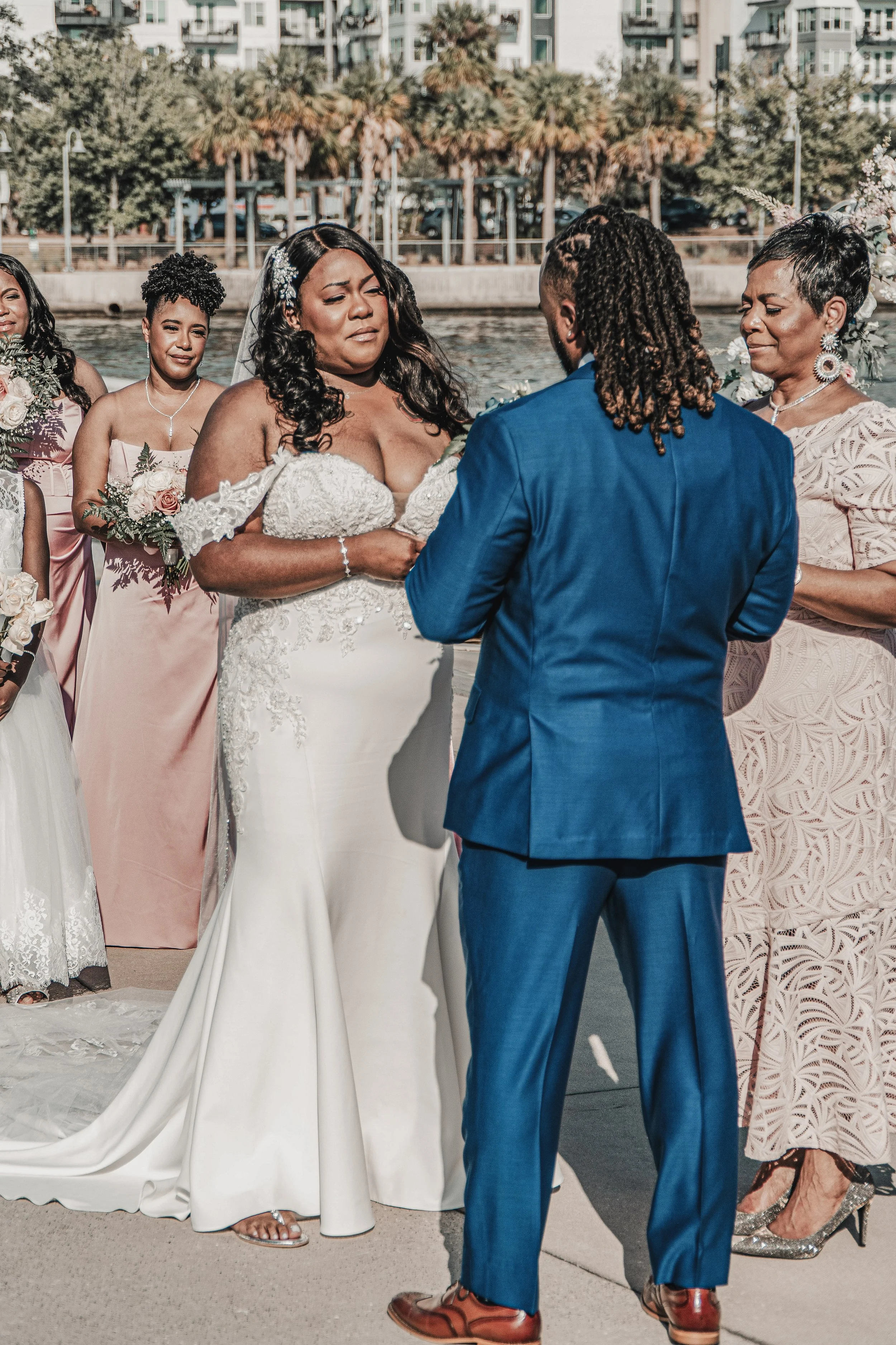 A wedding ceremony taking place outdoors near a body of water. The bride, wearing a white lace wedding gown, is standing next to a man in a blue suit, likely the groom. Bridesmaids and other women in formal dresses are surrounding them, with some hol