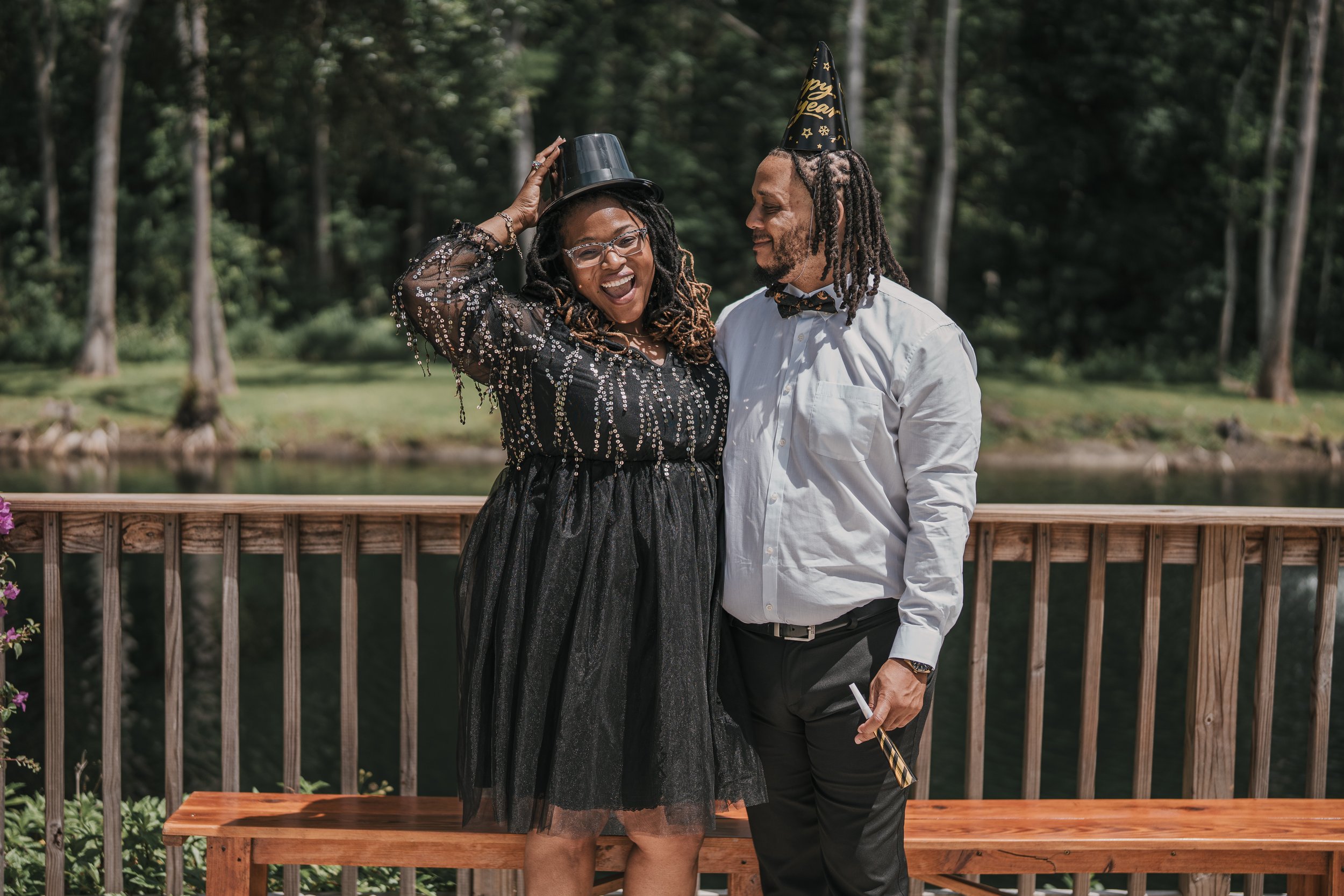 A woman in a black sequin dress and glasses, and a man in a white shirt with a checkered bow tie, celebrating New Year's Eve outdoors. They are wearing party hats and smiling, with the woman holding a black top hat and the man holding a party blowout