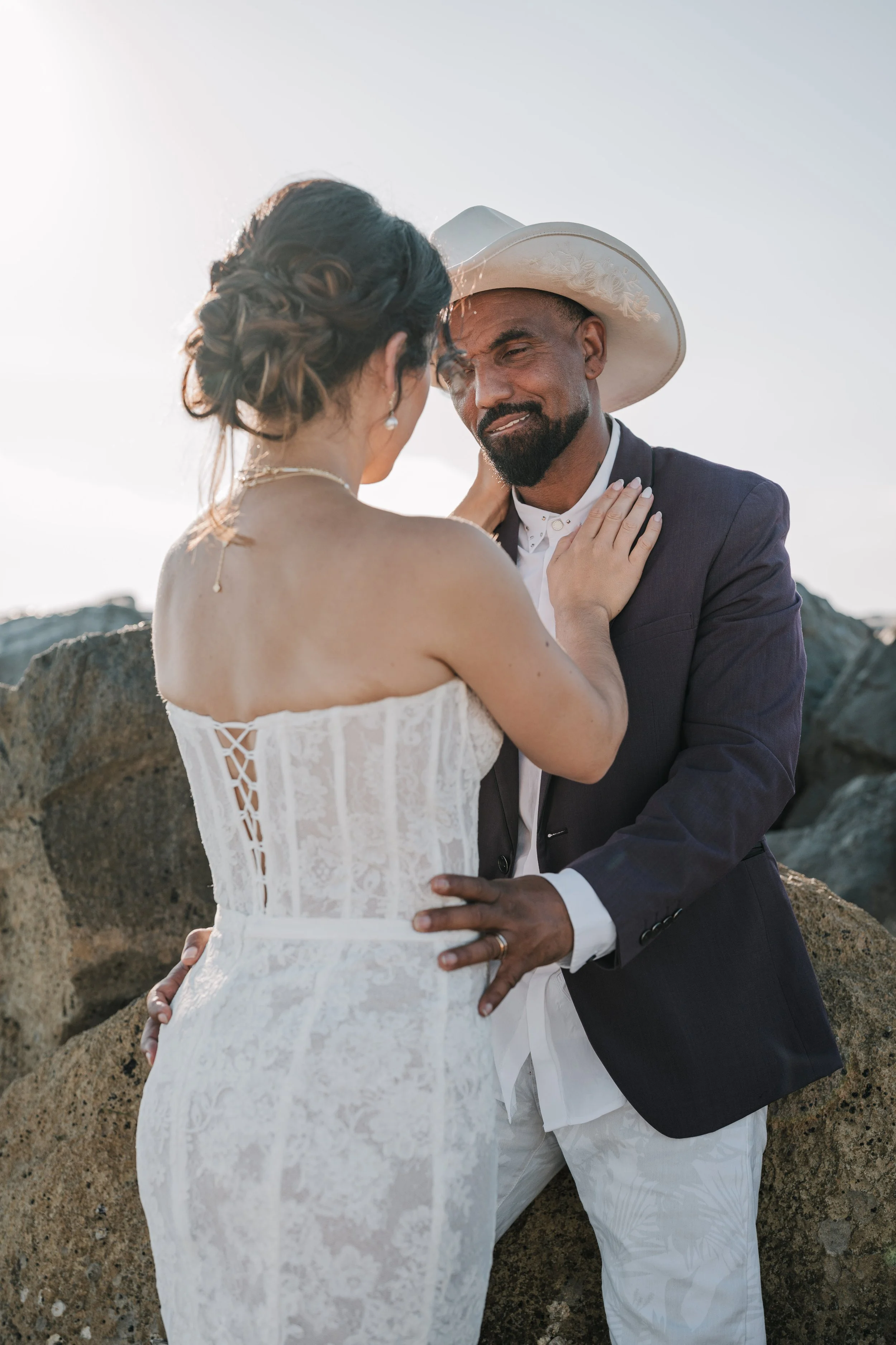 A couple dressed in wedding attire, standing close together outdoors on rocks, sharing an intimate moment at sunset with clear sky in the background.