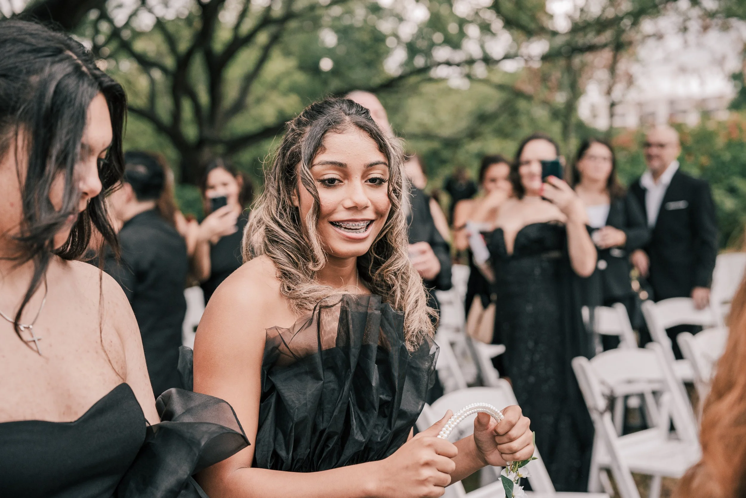 Young woman with curly hair and braces wearing a black dress at outdoor event, surrounded by people taking photos and sitting on white chairs.