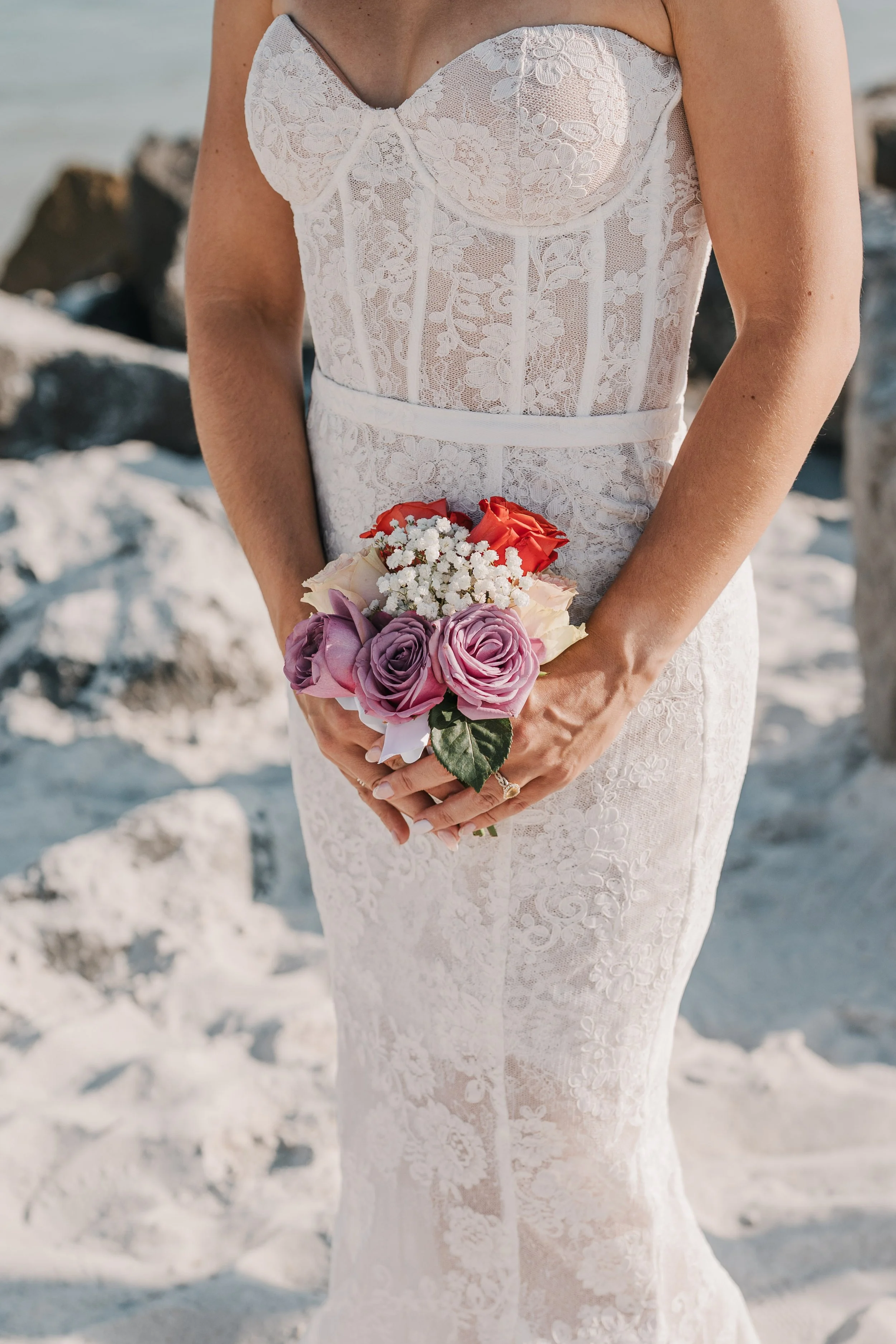 Close-up of a bride wearing a white lace wedding dress, holding a small bouquet of flowers with pink, red, and white roses and baby's breath, on a beach with rocks in the background.