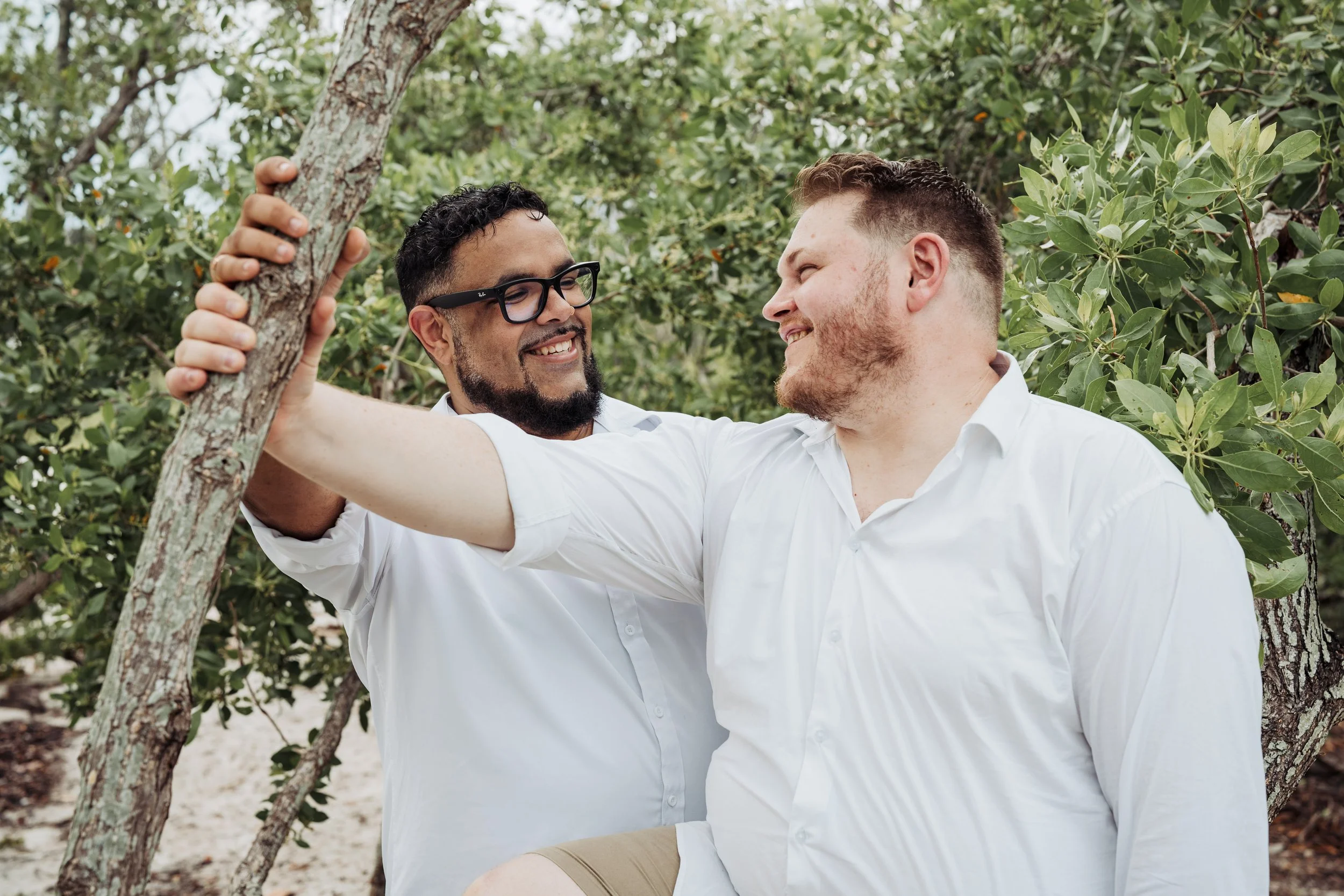 Two men smiling and looking at each other while standing outdoors among green bushes and trees. One man has glasses and a beard, and the other has short hair and a beard. They appear to be enjoying a moment together.