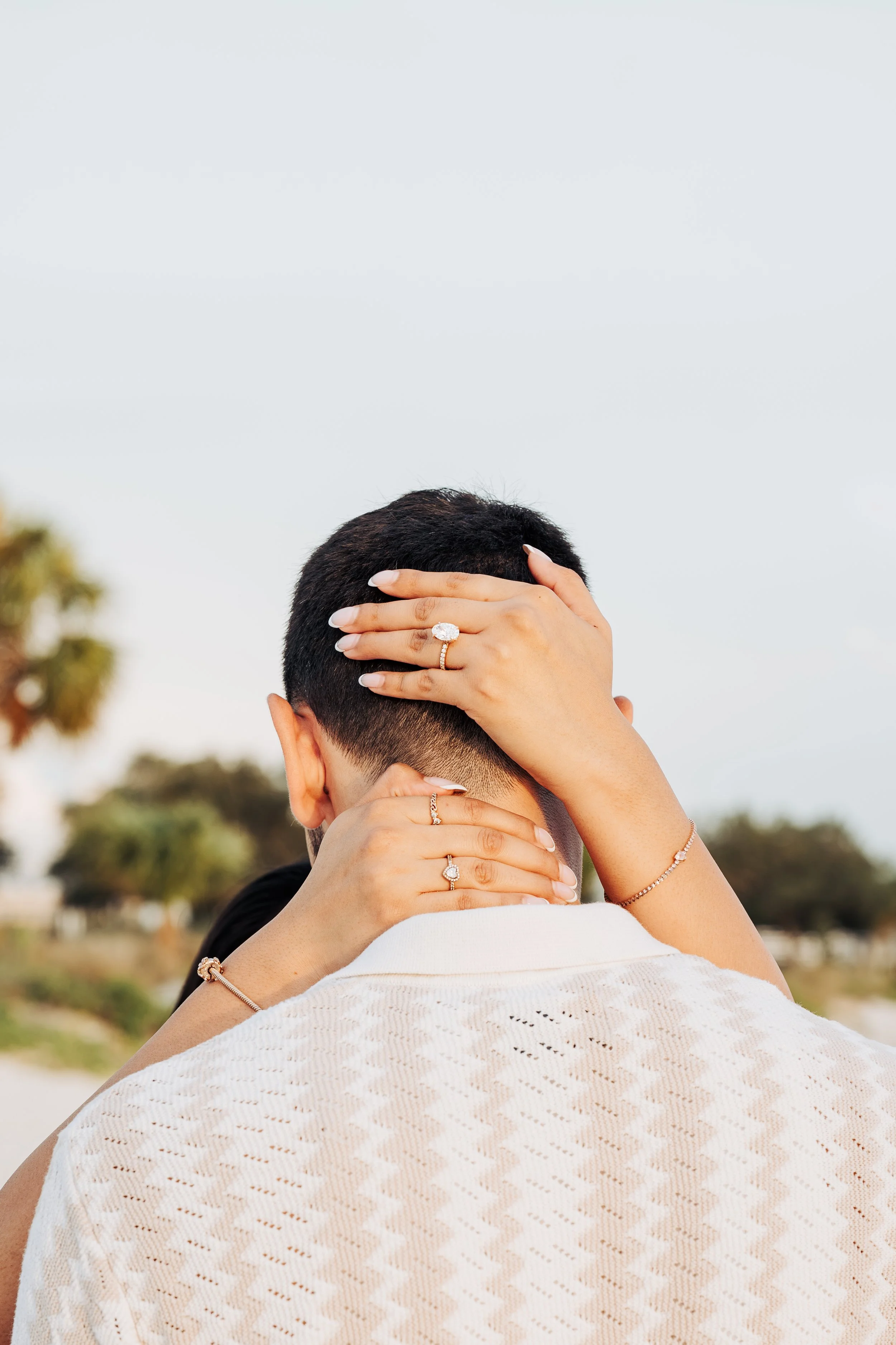 Close-up of four hands with rings and bracelets on their necks, touching the back of a person's head, outdoors with blurred trees and sky in the background.