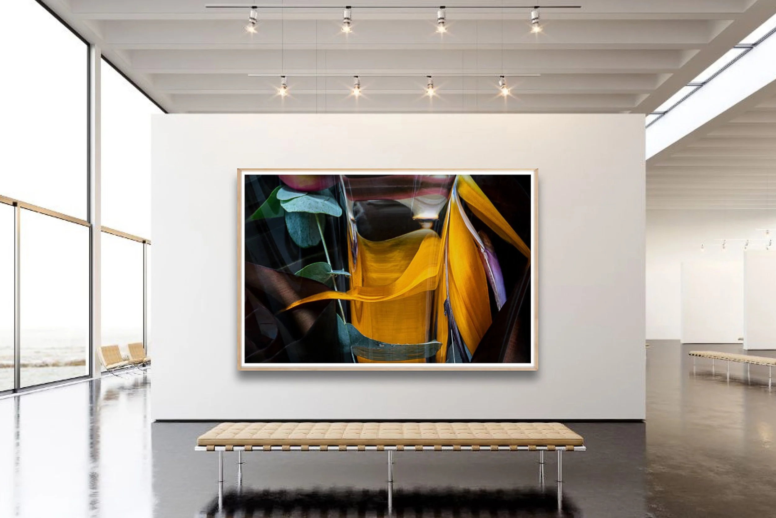 An art gallery with a large colorful abstract painting displayed on a white wall, with natural light coming through large glass windows on the left, a bench in front, and a ceiling with track lighting.