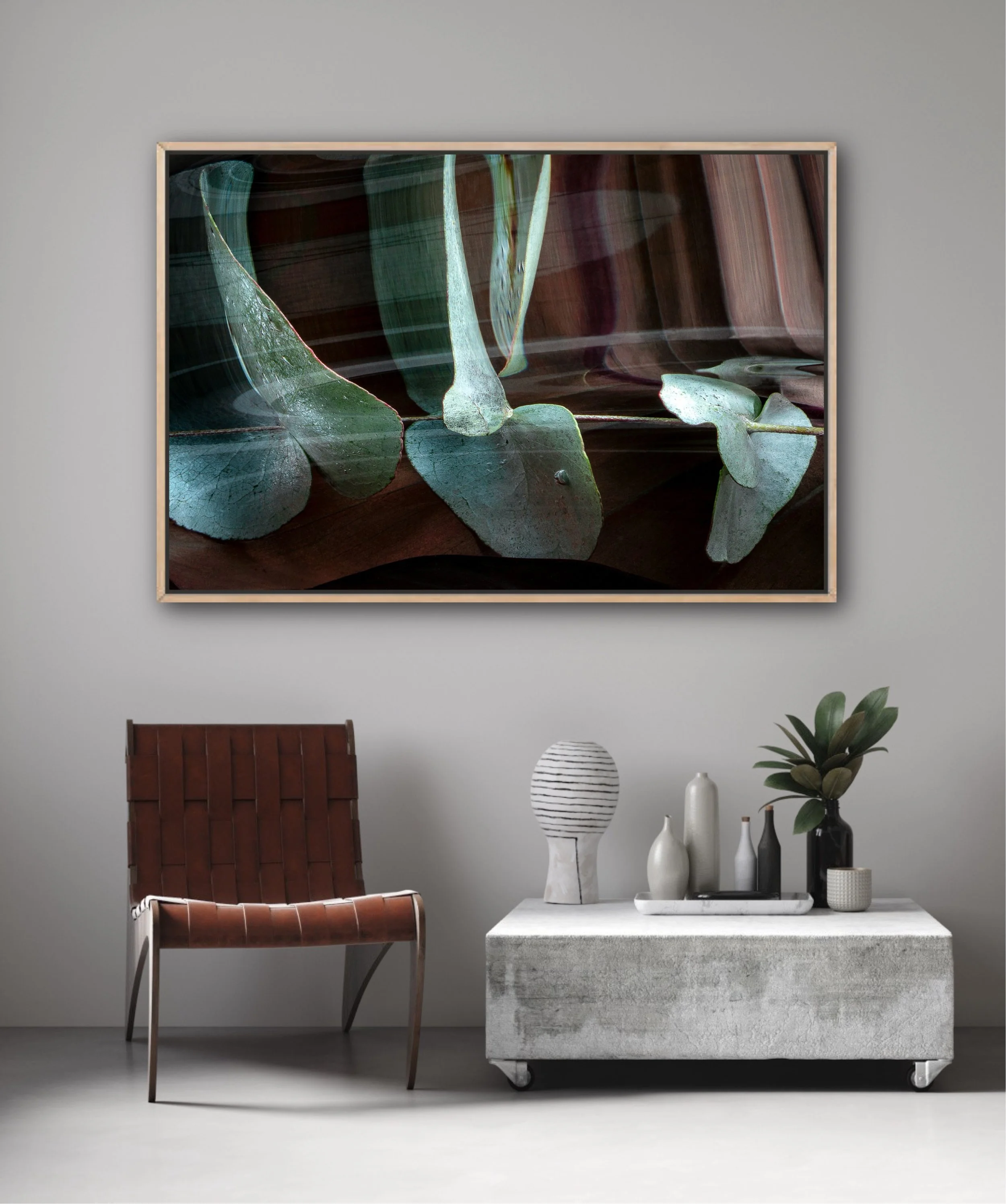 A modern interior scene with a large framed abstract artwork of green leaves on a gray wall. In front, there is a brown leather chair, a white marble table on wheels with decorative white and black vases, and a small potted plant.