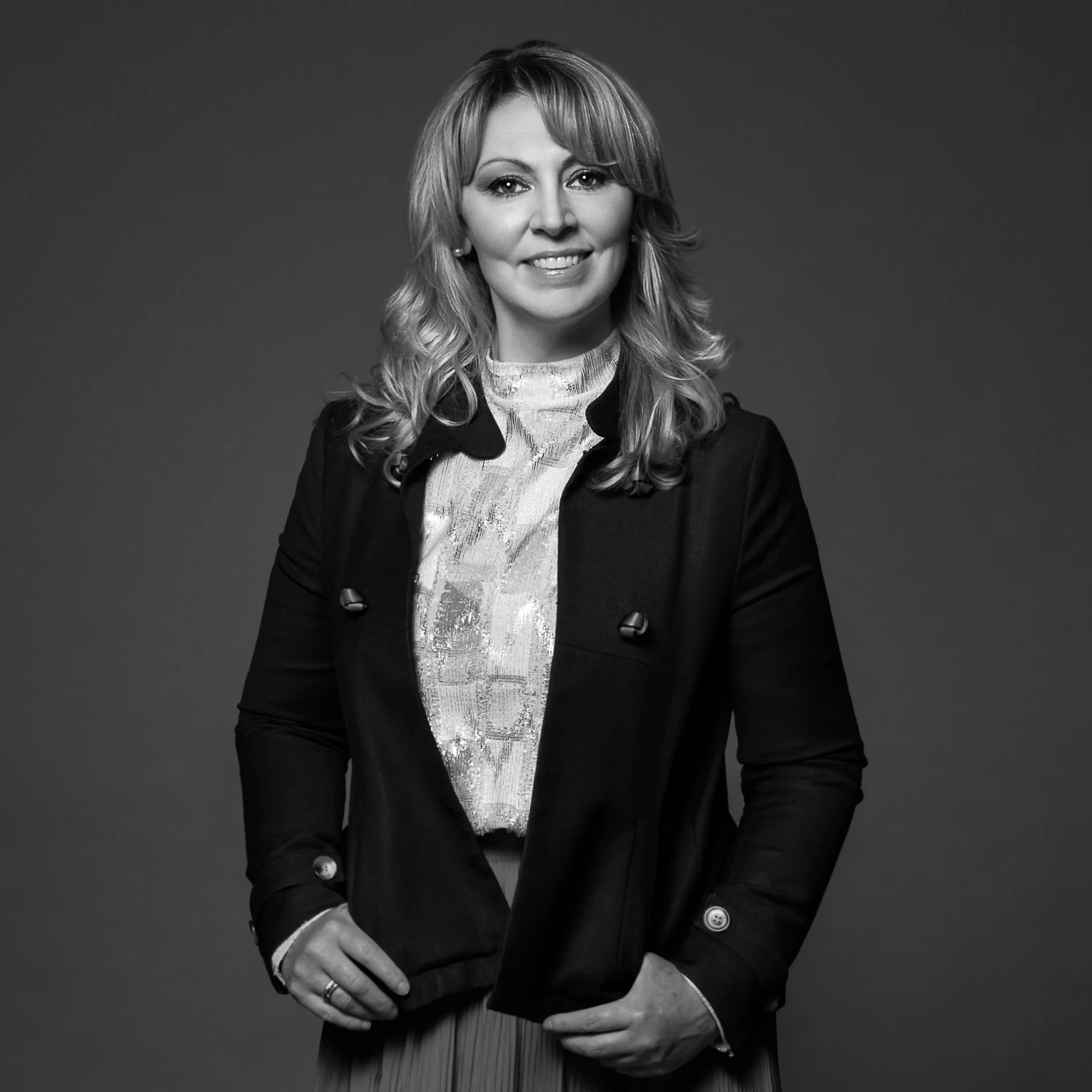 Black and white portrait of a woman with shoulder-length wavy hair, wearing a blazer and a patterned blouse, smiling with her hand resting on her waist.