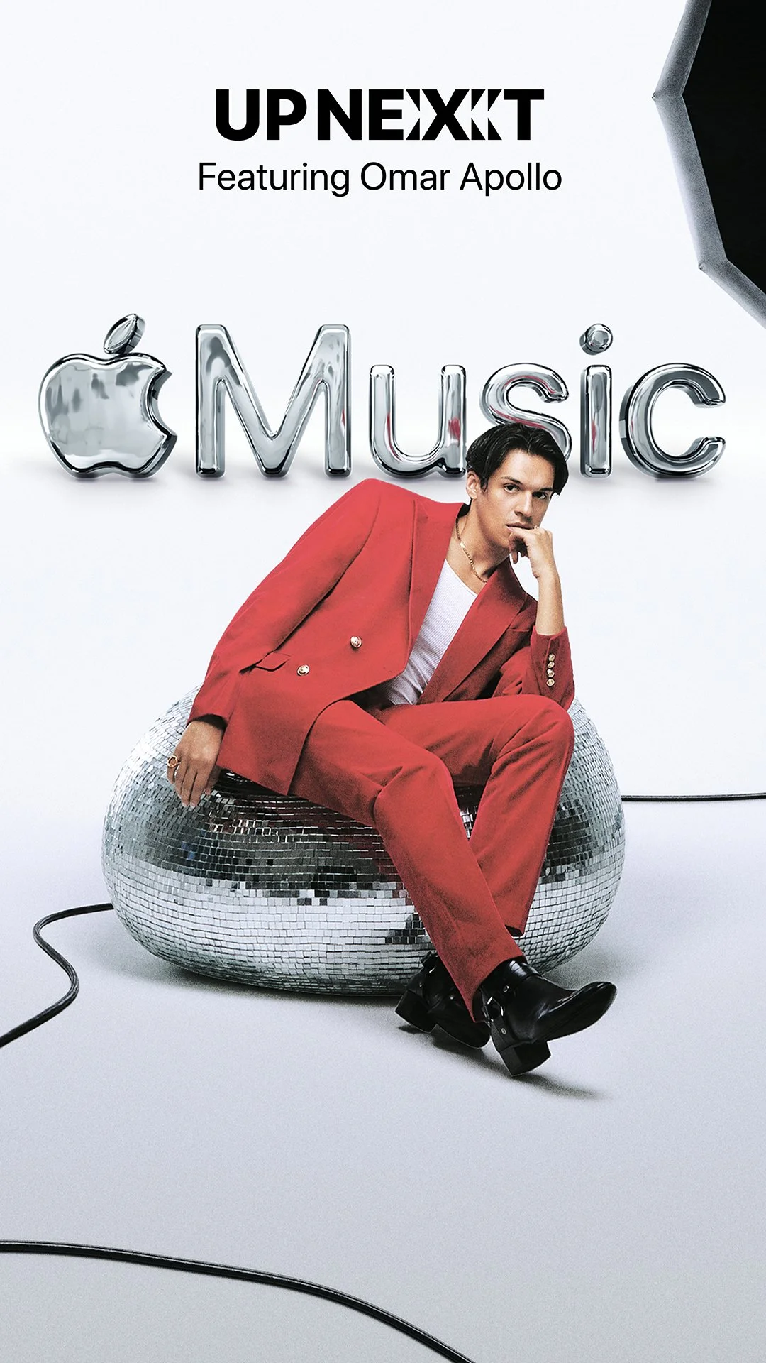 Person sitting on a mirrored disco ball in front of a backdrop with the Apple Music logo and the text 'Up Next featuring Omar Apollo'.