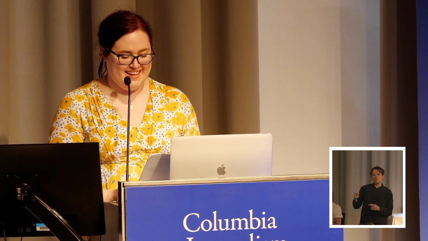 A woman with glasses and red hair, wearing a yellow floral dress, is speaking at a podium with a blue sign that says Columbia. She is smiling and looking at a laptop in front of her.