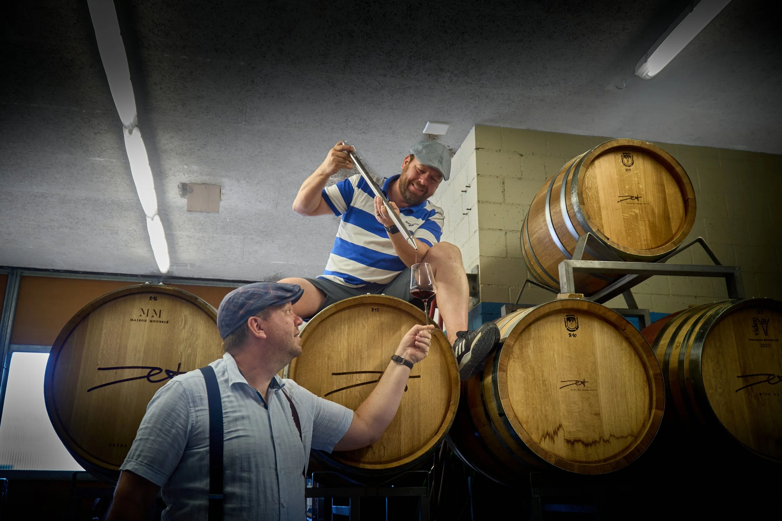 Zwei Männer in einem Weinkeller, einer sitzt auf einem Weinfass und füllt einem anderen eine Glass Wein. Der Mann auf dem Fass trägt ein gestreiftes T-Shirt und eine Mütze, der andere einen Hut und ein Hemd mit Hosenträgern.