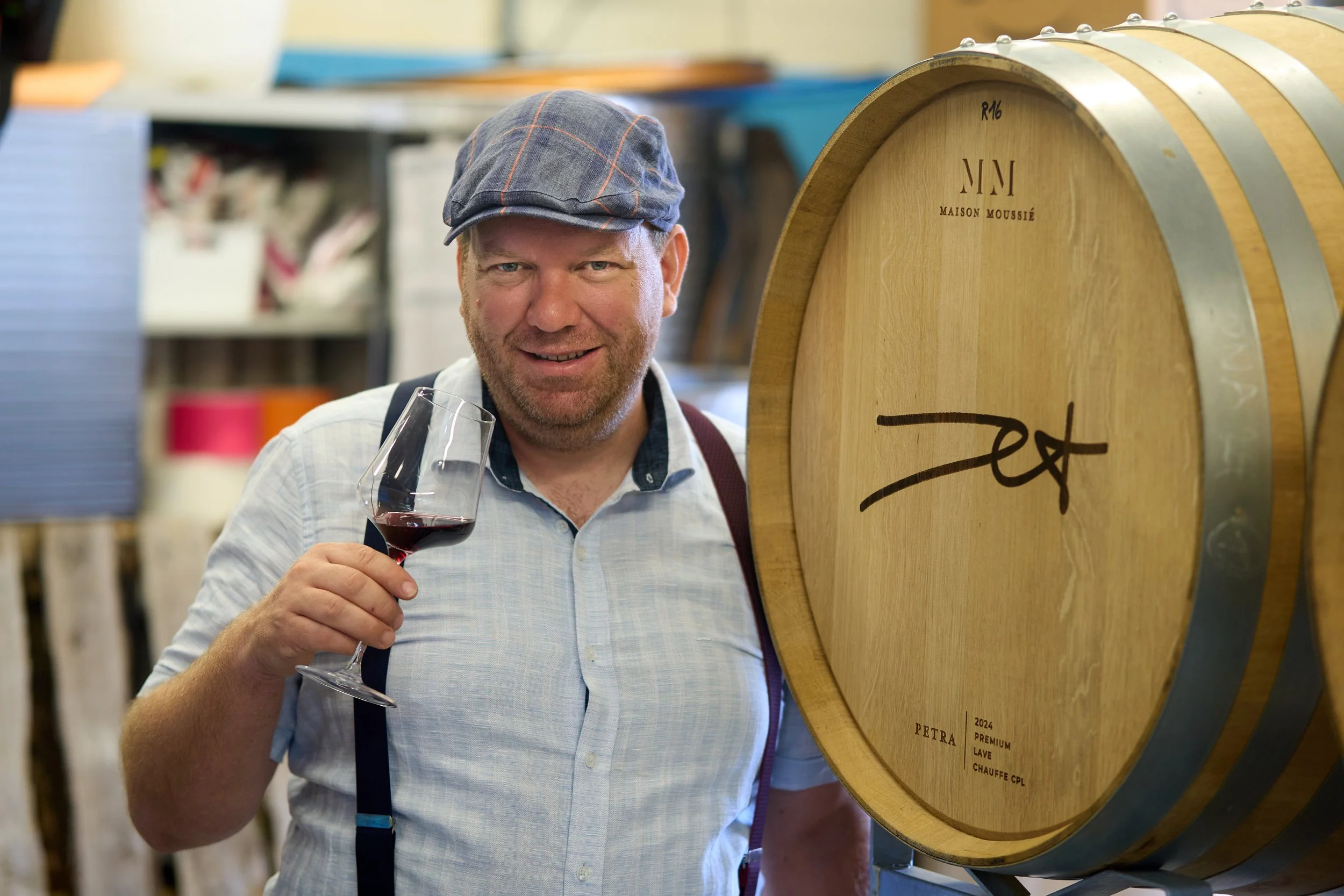 Ein Mann in Hemd und Kappe hält ein Glas roten Wein in der Hand vor einer Wein- oder Spirituosenlagerung