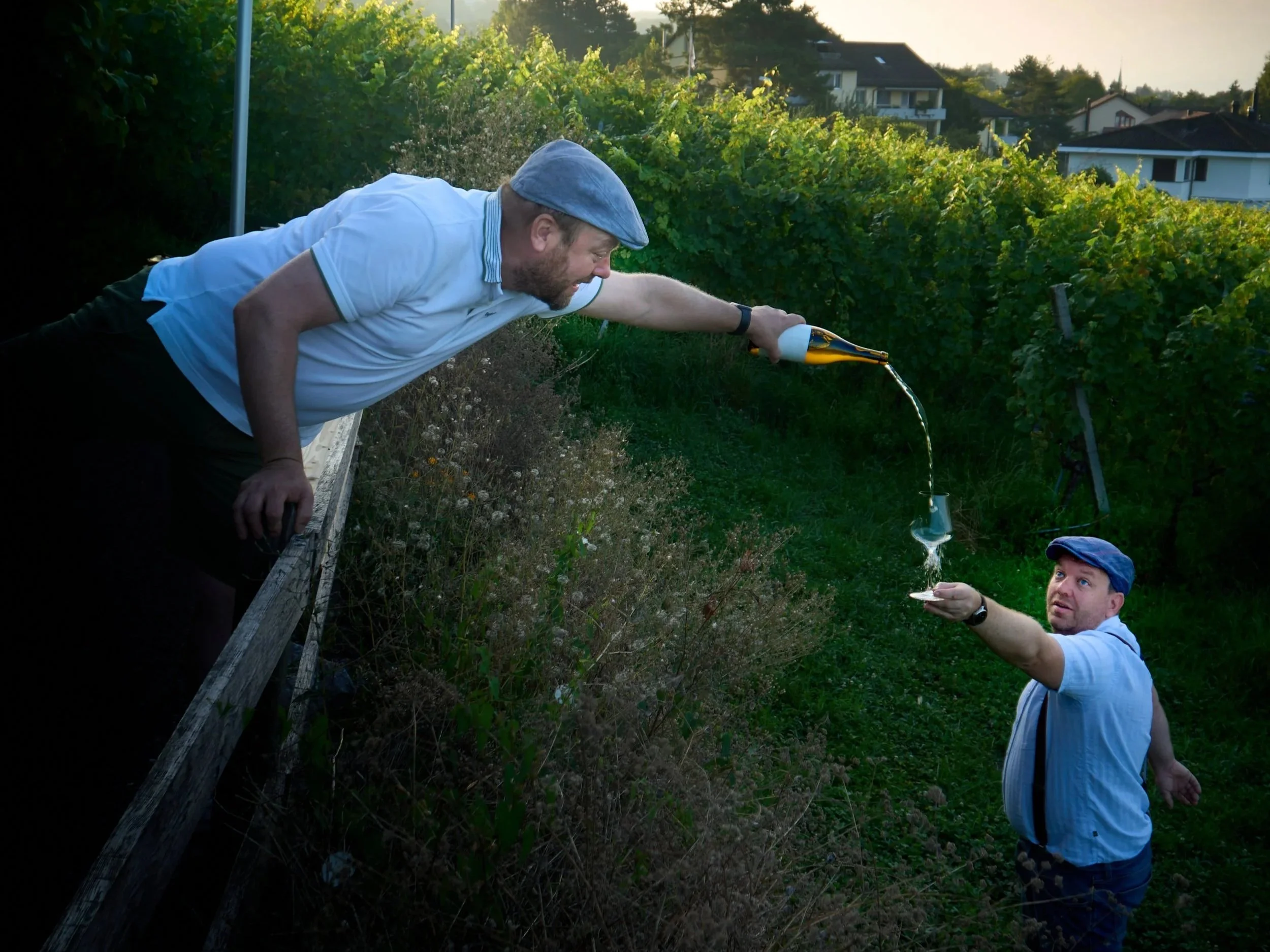 Zwei Männer im Garten, einer sitzt auf einem Zaun und hält ein Glas, woraus ein Getränk in ein anderes Glas gegossen wird, während der andere Mann im Stehen Wasser oder Sekt eingießt. Es ist Abend, und die Männer tragen Hüte und Kurzarmhemden.