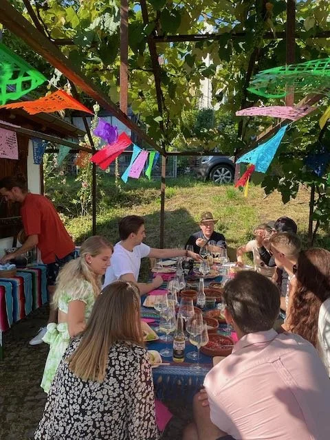 Menschen sitzen an einem langen Tisch im Freien und genießen eine Mahlzeit. Der Tisch ist mit bunten Tischdecken gedeckt und es hängen dekorative Girlanden über ihnen. Es scheint ein sonniger Tag zu sein und die Umgebung ist grün und gemütlich.