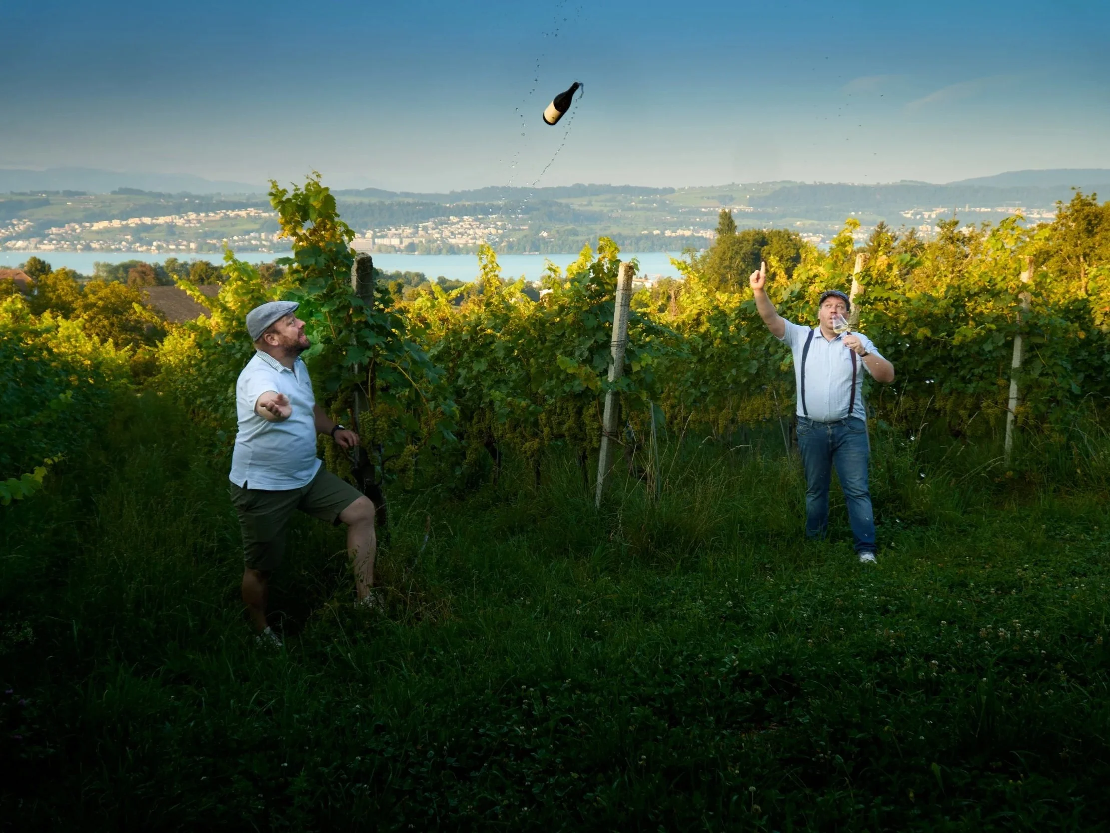 Zwei Männer in einem Weingarten, einer schlägt gegen einen reifen Weintraubenzweig, der andere wirft eine Weinflasche in die Luft, im Hintergrund ein Fluss und Berge.