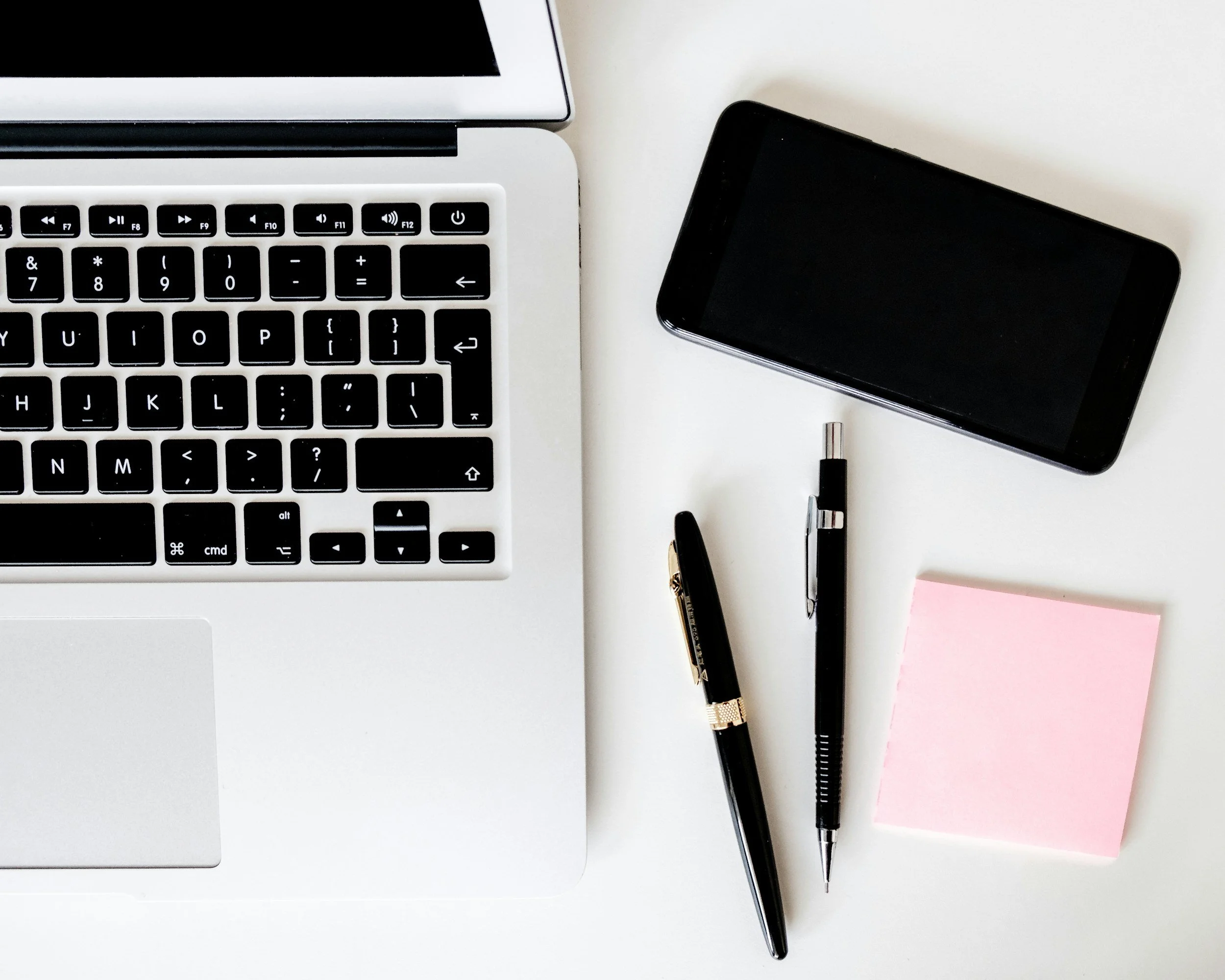 Top view of a workspace with a silver laptop, a black smartphone, two black pens, a pink sticky note, on a white surface.