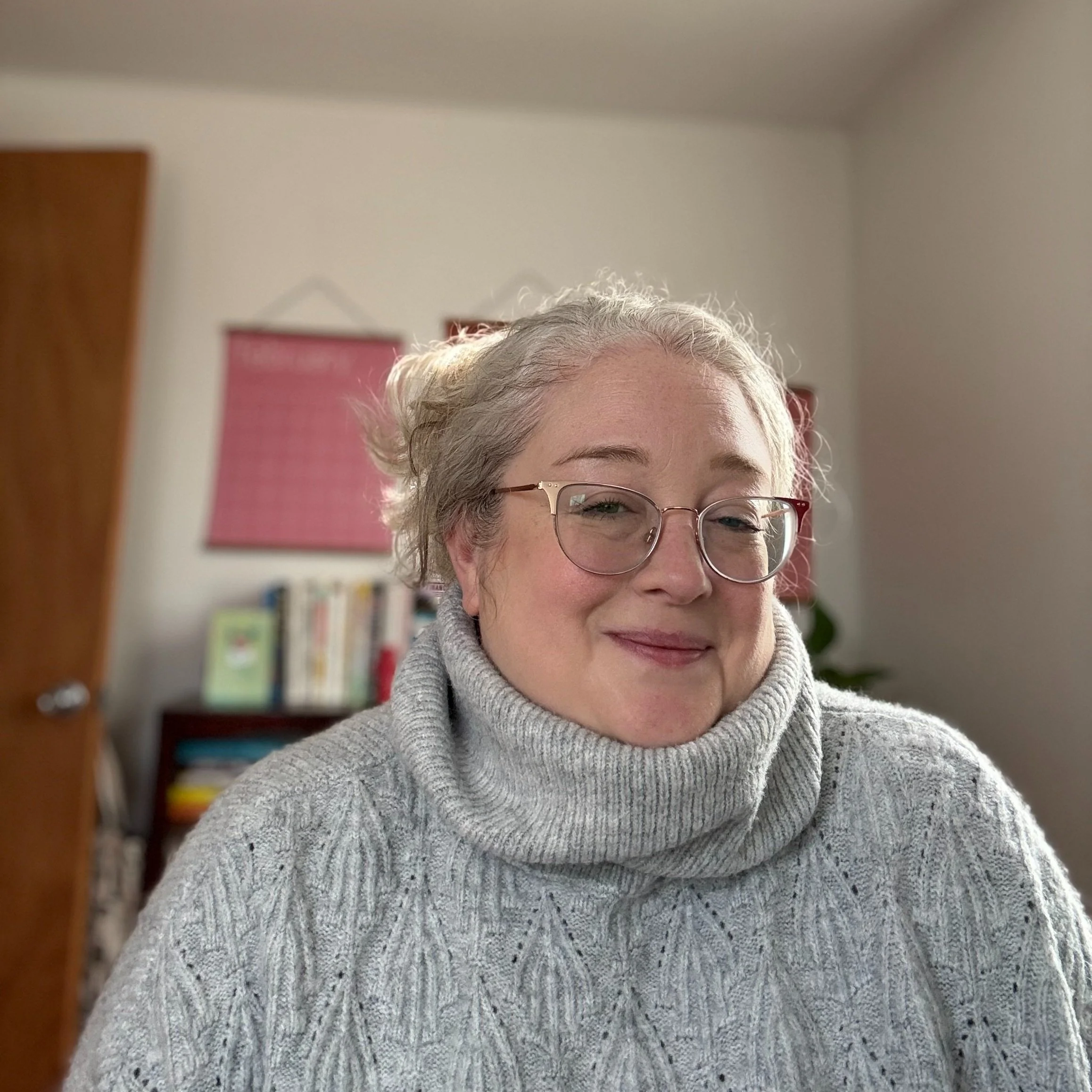 A woman with glasses and gray hair styled in a bun, wearing a gray cable-knit sweater with a turtleneck, smiling at the camera inside a room with a bookshelf and artwork in the background.