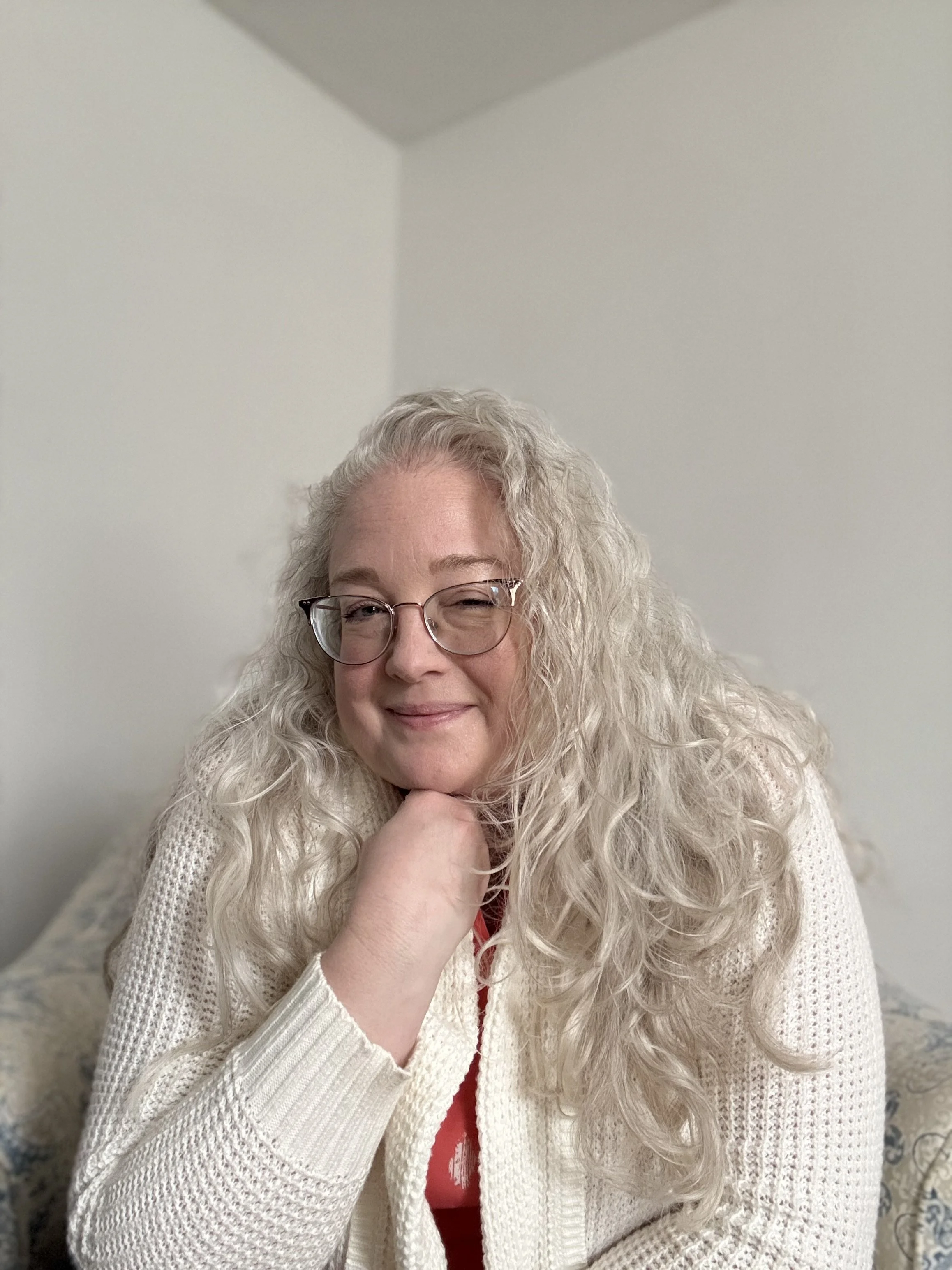 A woman with long, curly blonde hair and glasses sitting on a couch, resting her chin on her hand, smiling at the camera.