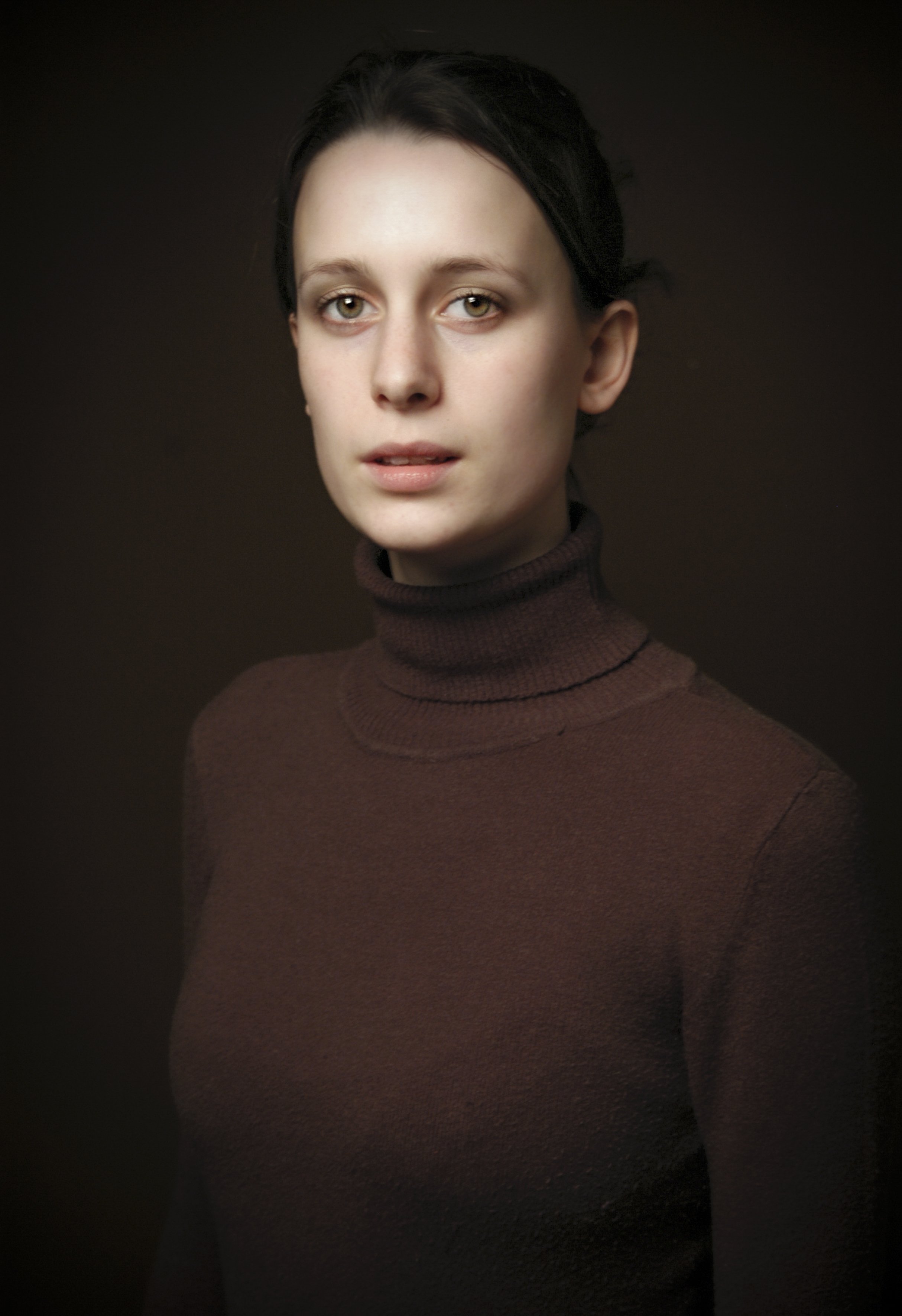 Portret van een jonge vrouw met donker haar, lichtgekleurde ogen en een bruin gebreid turtleneck shirt, tegen een donkere achtergrond.
