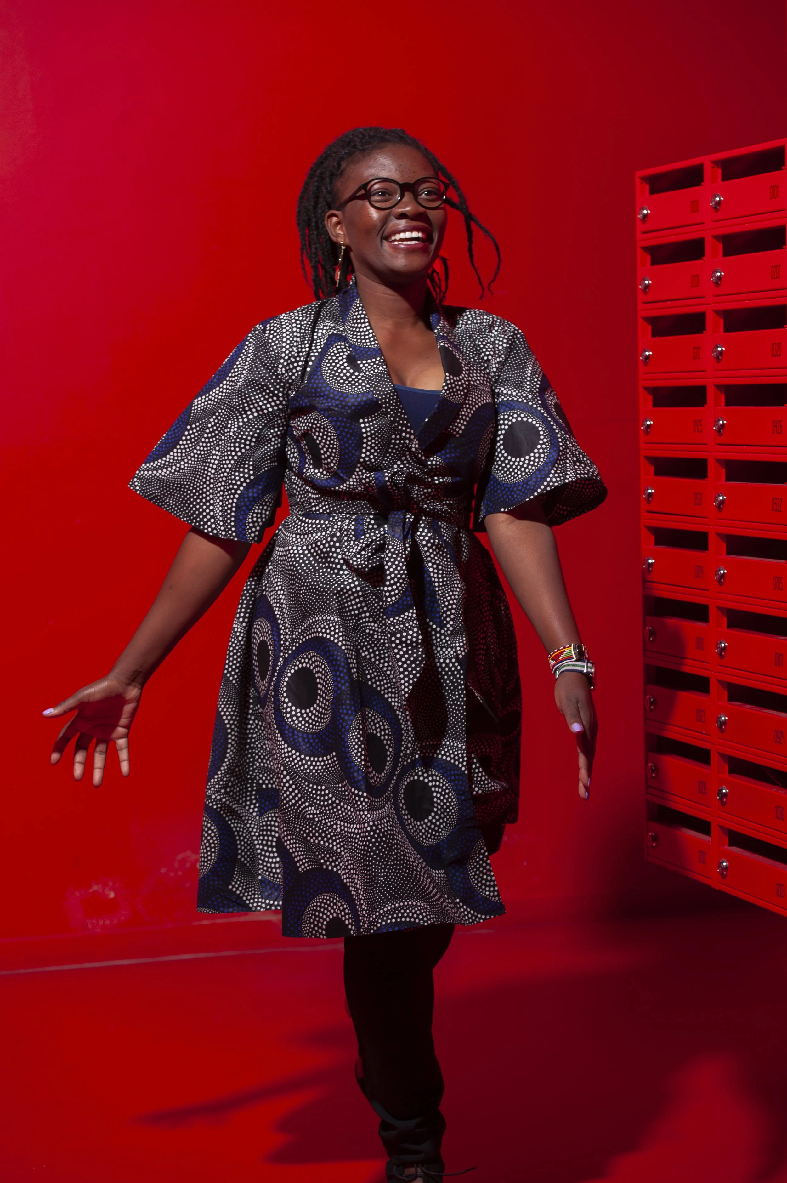 Een lachende vrouw met dreadlocks, gekleed in een zwart-wit patroon jurk, staat voor een rode muur en een bank met postvakjes.