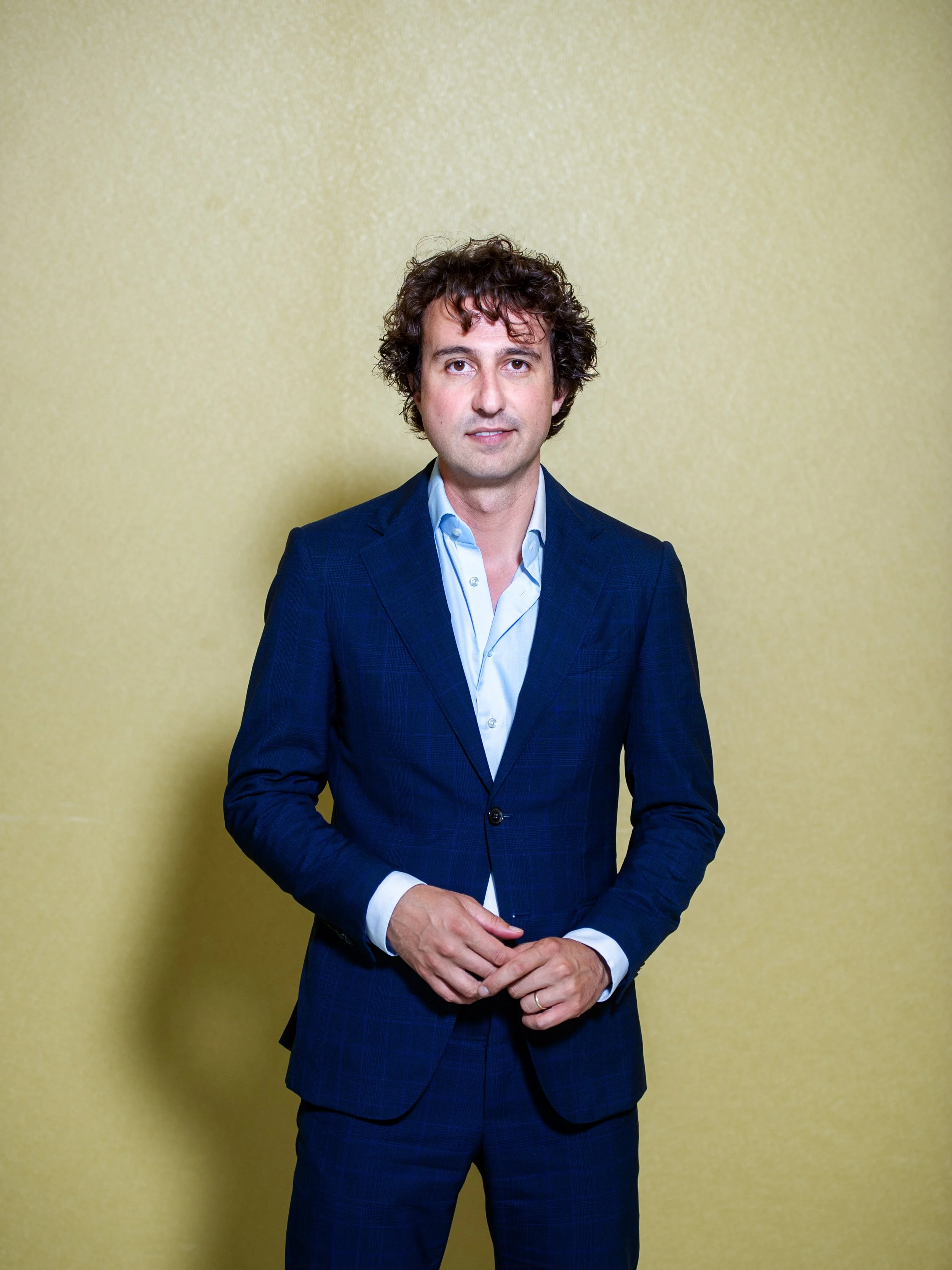 Man in een blauw pak met witte overhemd, staand voor een gele muur, kijkend naar de camera.