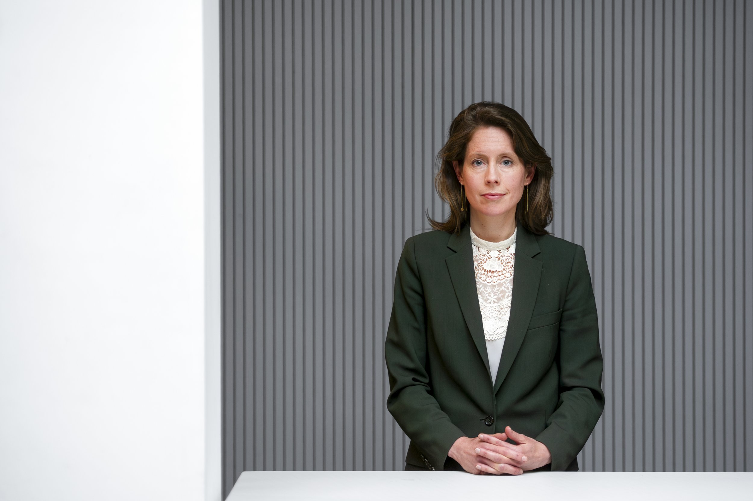 Portret van een vrouw in een zakelijke outfit die recht naar de camera kijkt, zittend aan een tafel, met een achtergrond van verticale grijze lijnen.