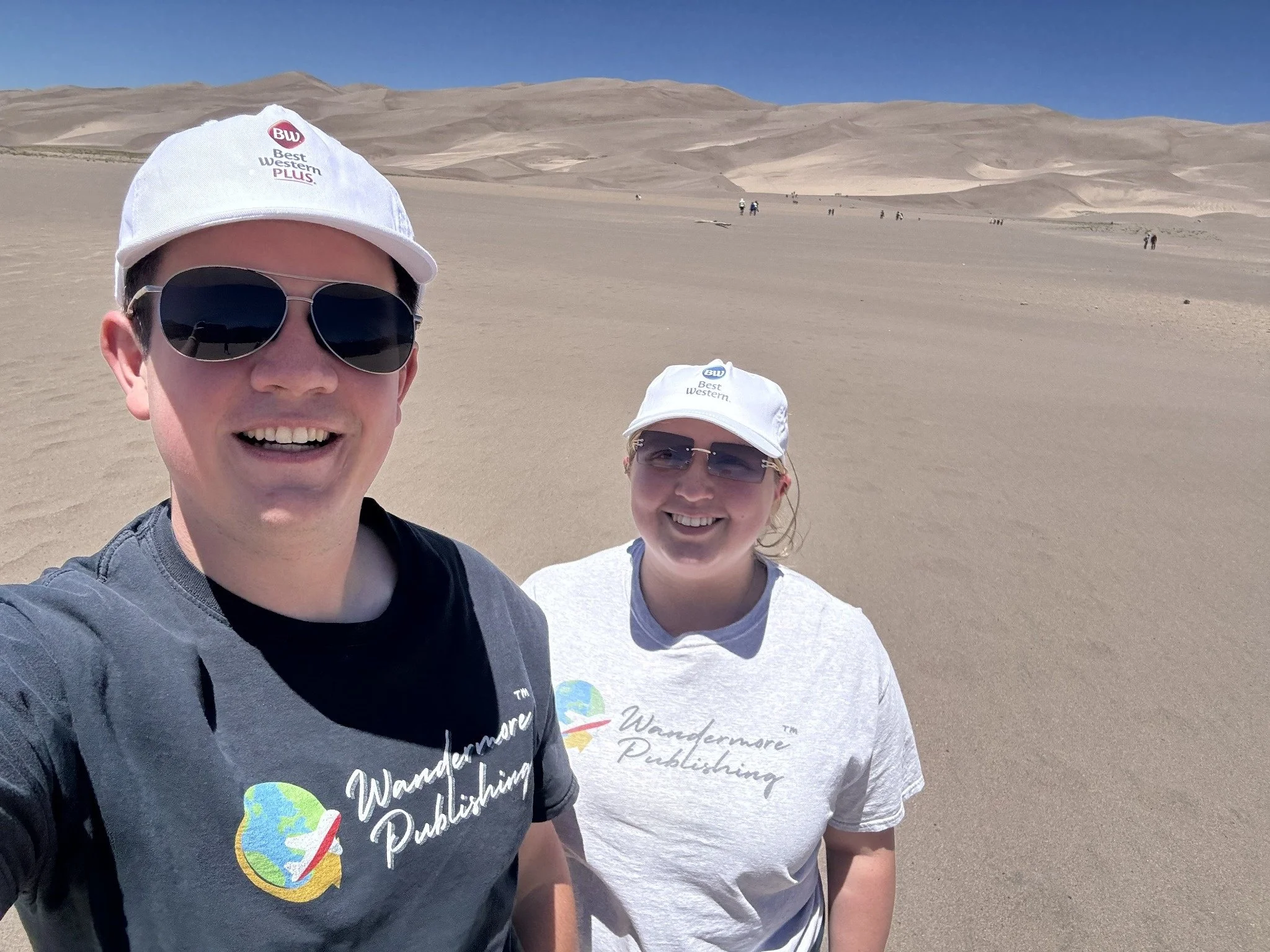 Two people standing in a desert landscape with dunes in the background, both wearing white caps and holding the camera to take a selfie.
