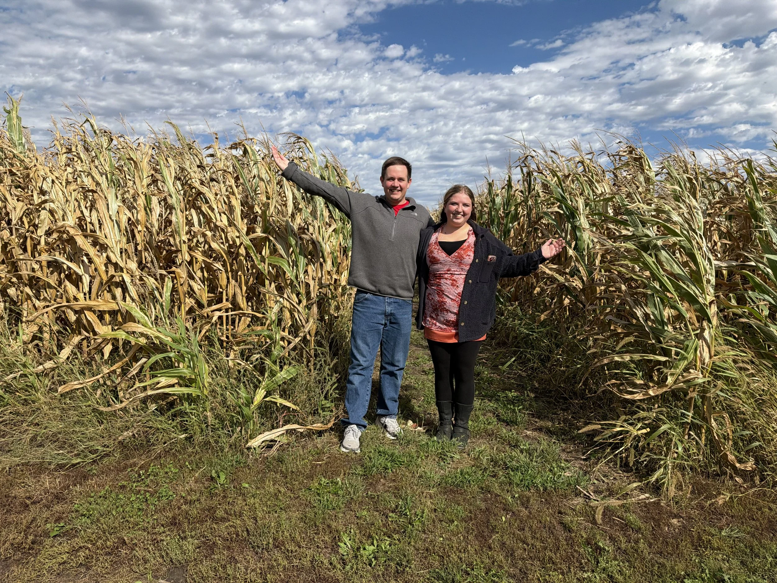 Two smiling people, a man and a woman, standing in a soybean field under a partly cloudy sky. The man has his arm raised and is wearing a gray shirt, while the woman is wearing a black jacket and a patterned shirt.