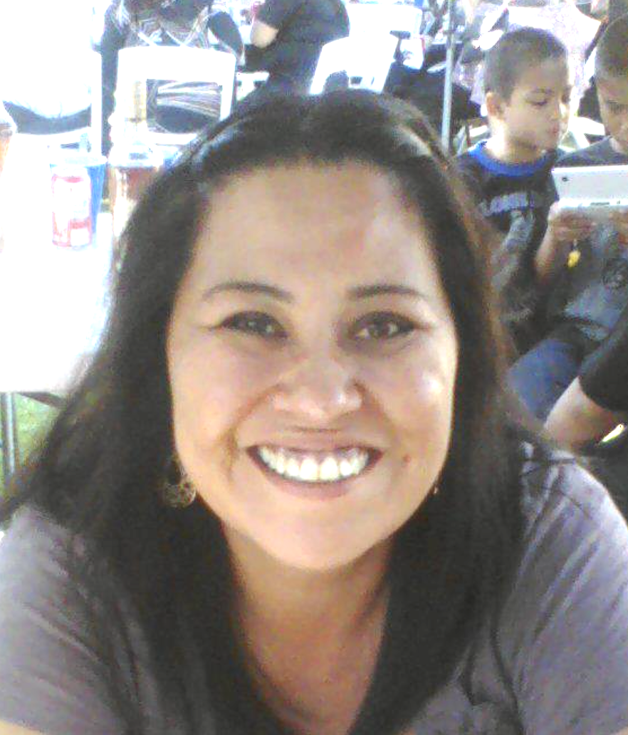 Smiling person at a social gathering sitting at a table with drinks, with children in the background playing on electronic devices.