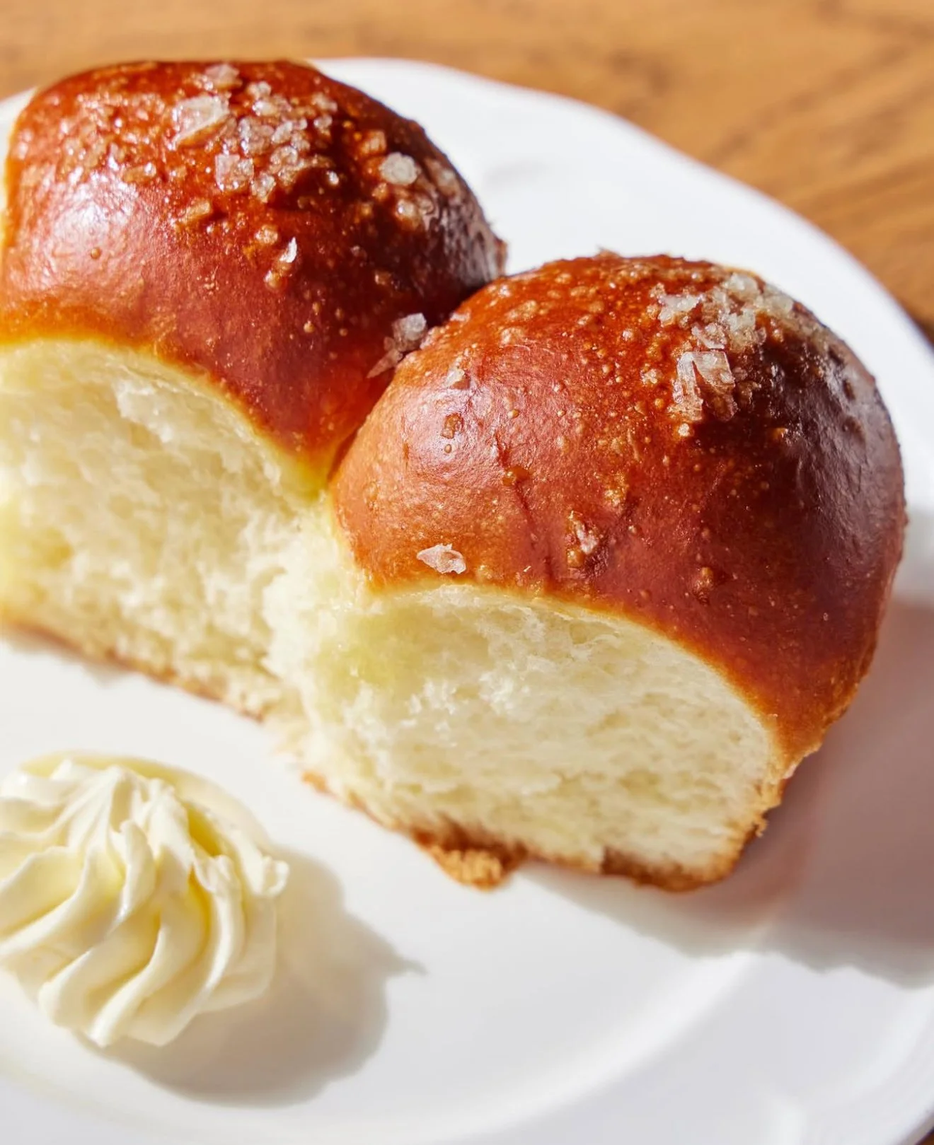 Close-up of two bread rolls with a golden-brown crust sprinkled with coarse salt, served on a white plate with a dollop of butter.