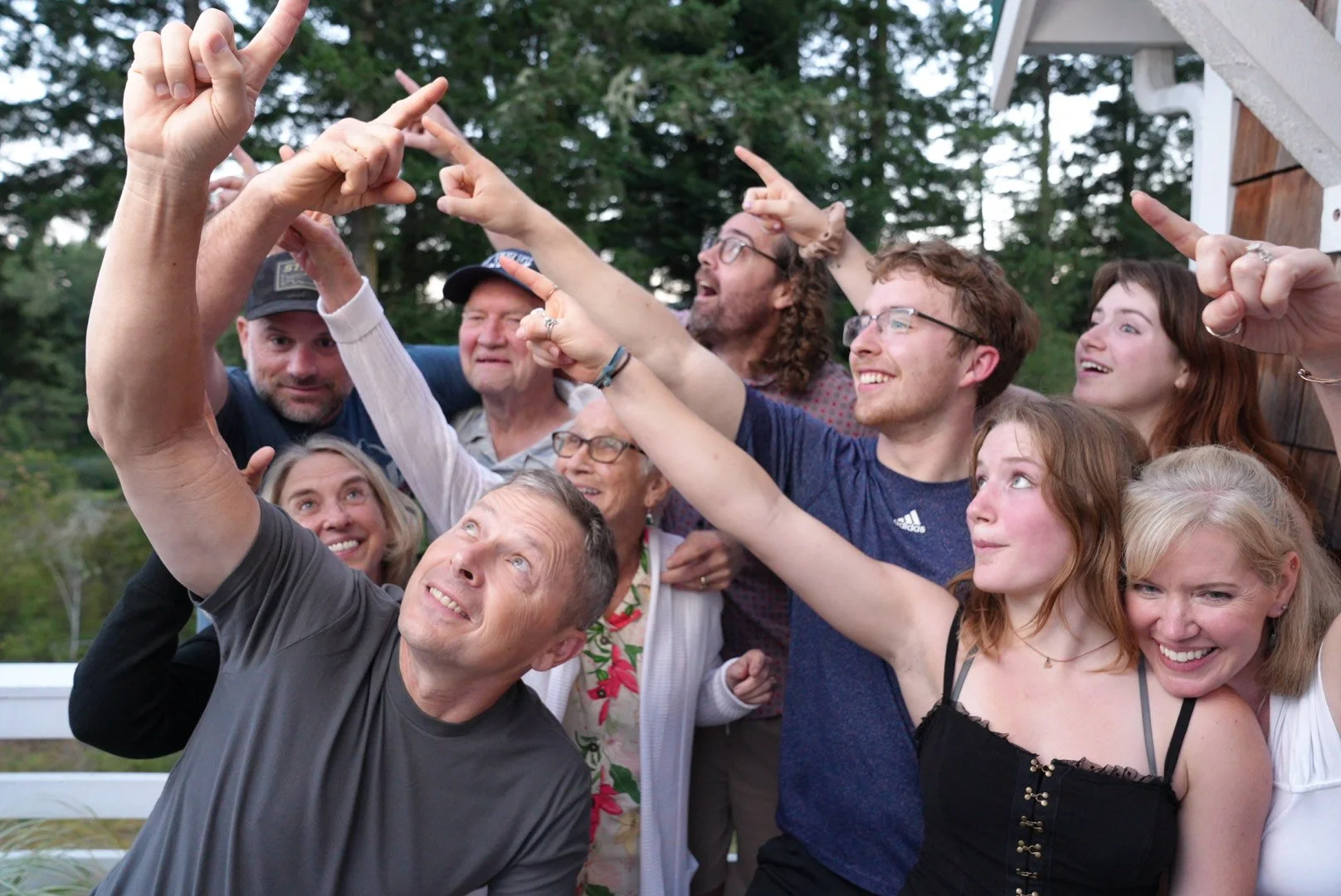 Group of people taking a selfie outdoors with greenery in the background, smiling and pointing their fingers in different directions.