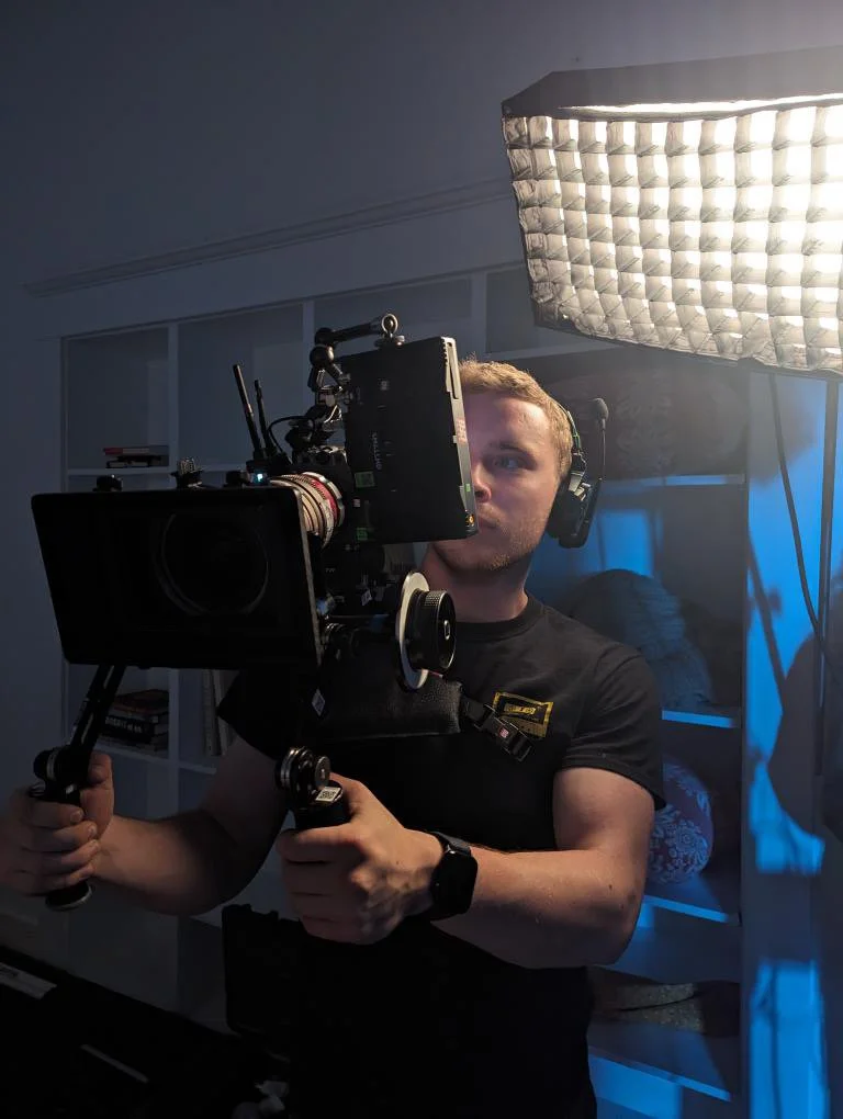 A young man operating a professional camera with a large light and softbox, wearing headphones and a black T-shirt in a studio setting.