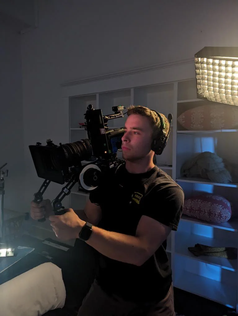 A man operating a professional video camera on a shoulder rig in a room with shelves and soft box lighting.