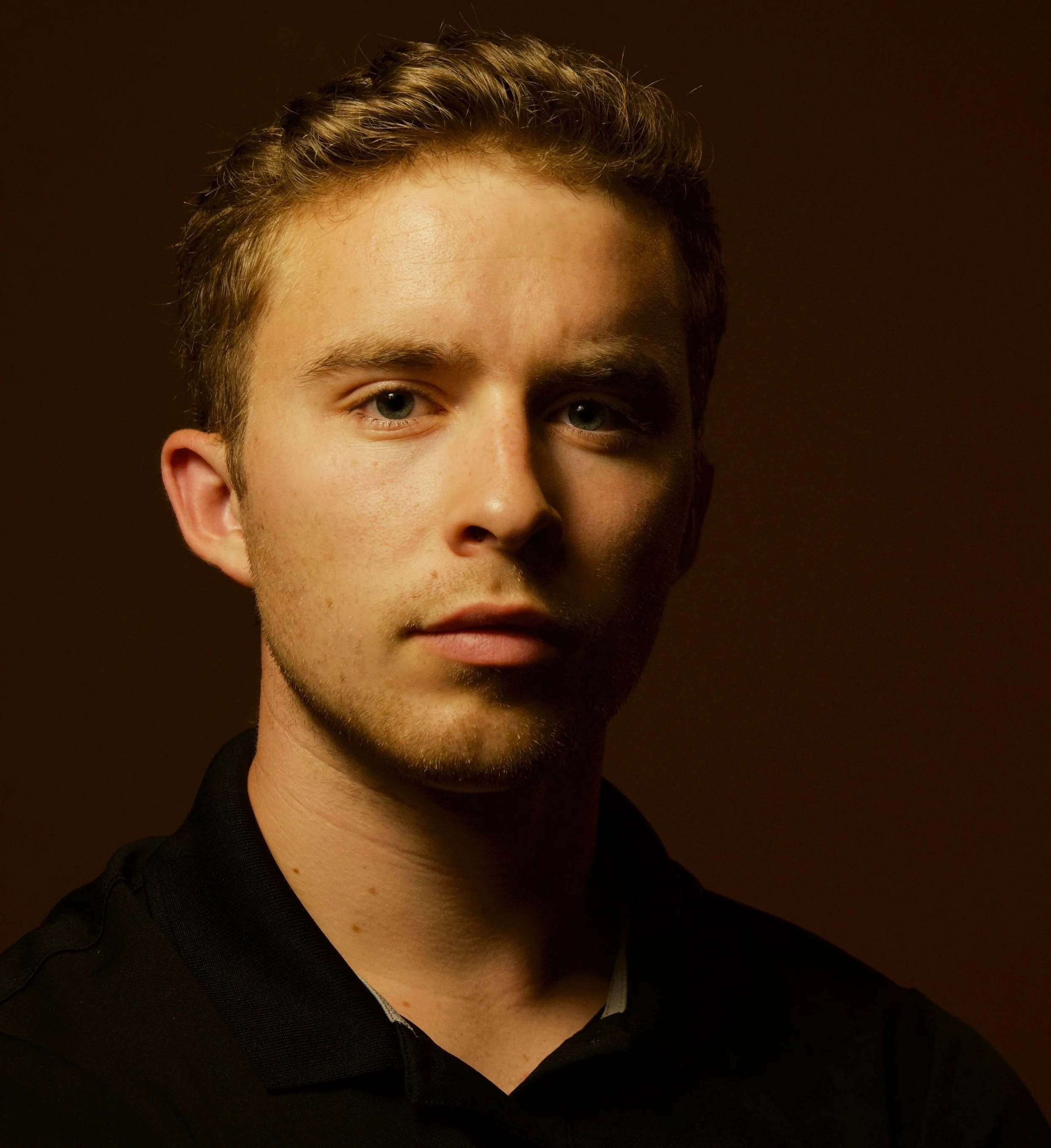 Close-up of a young man's face with bright blue eyes, short wavy hair, fair skin with freckles, wearing a black shirt against a dark background.