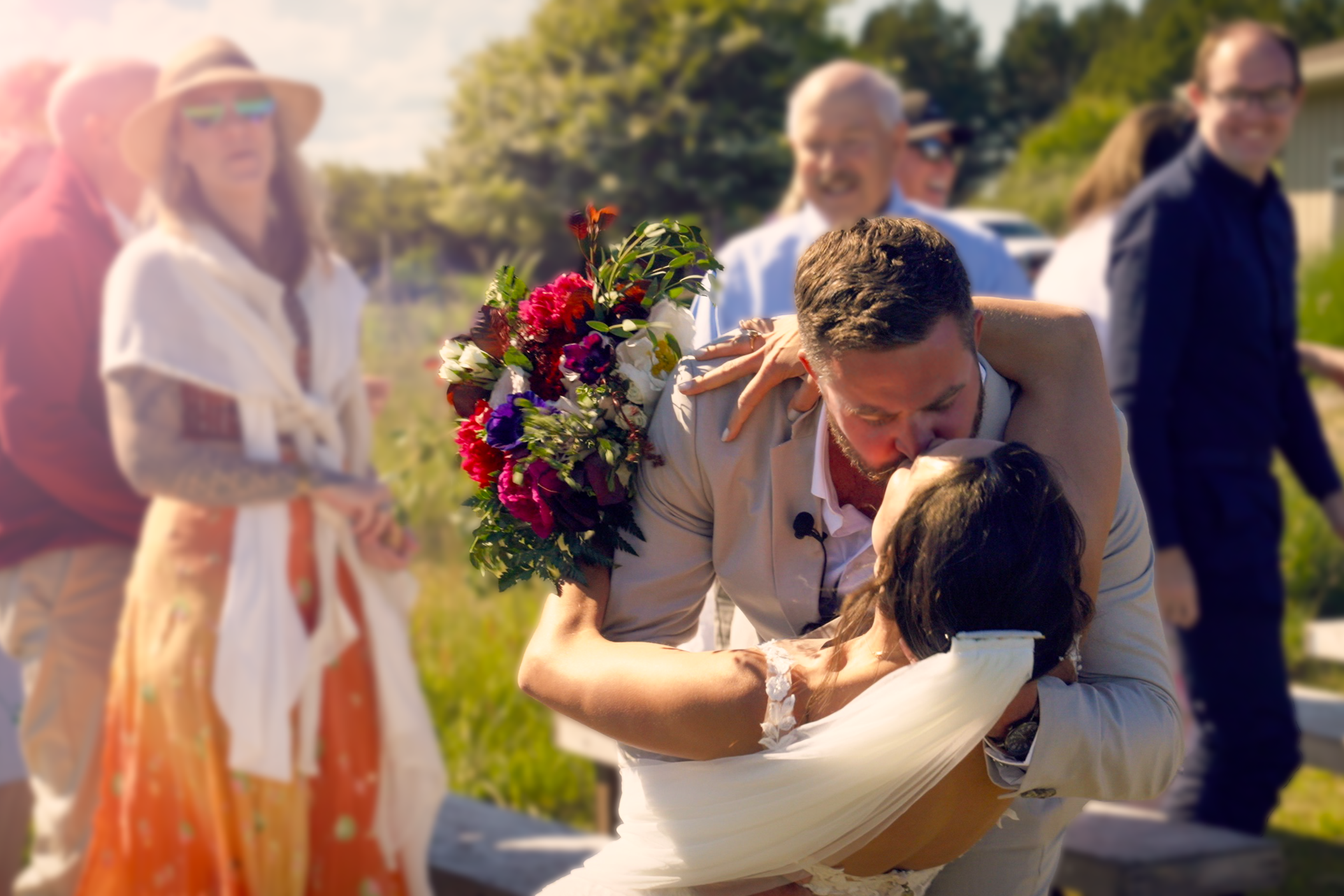People celebrating a wedding outdoors on a sunny day, with one man holding a bouquet of flowers and leaning in for a kiss with a woman, while others smile and watch in the background.
