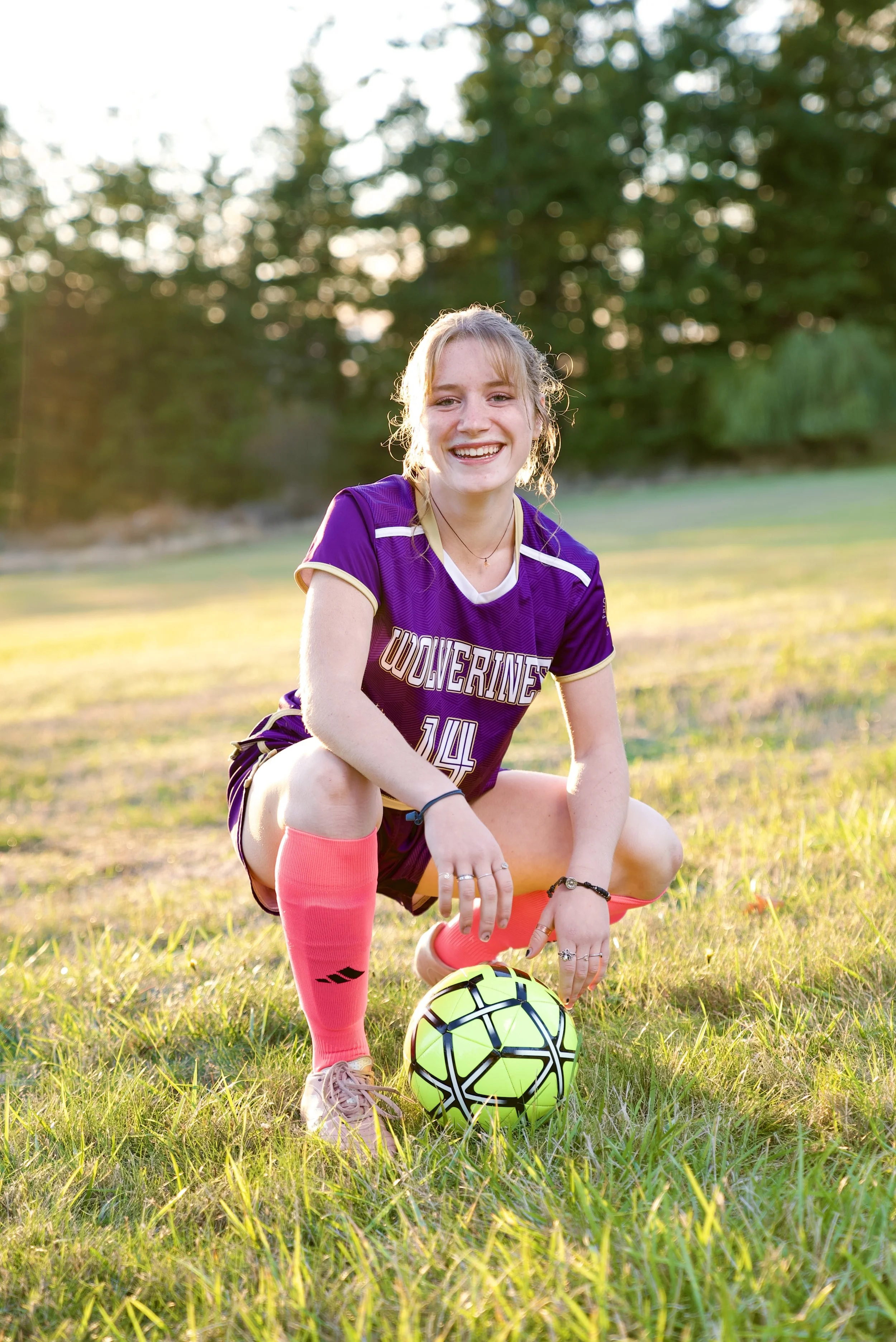 Young woman in a purple sports uniform with the word 'Wolverines' on it, kneeled on the grass with her left knee, holding a neon yellow-green and black soccer ball, outdoors on a sunny day with green trees in the background.