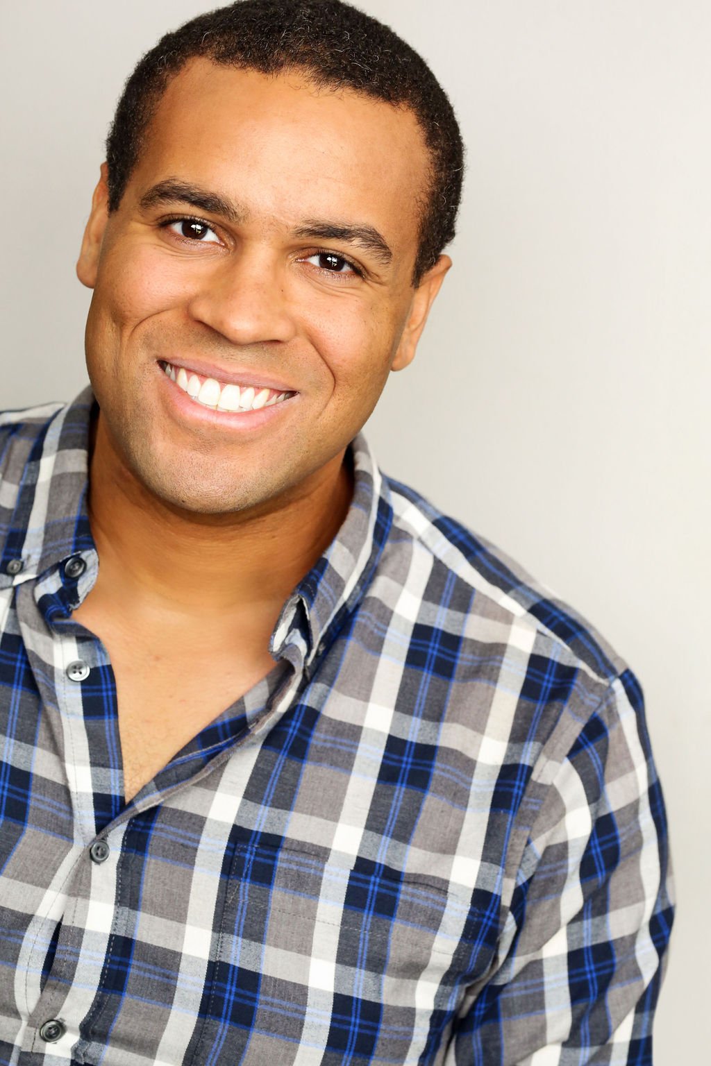 A smiling man with short dark curly hair wearing a gray, blue, and white plaid button-up shirt, standing against a plain light-colored background.