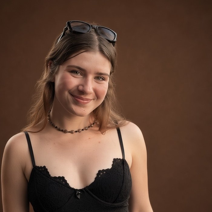 A young woman with shoulder-length light brown hair, wearing glasses on her head, a black lace camisole, and a beaded necklace, smiling in front of a brown background.