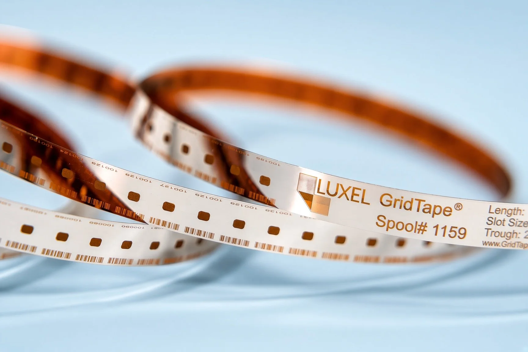 Close-up of a roll of LUXEL GridTape, a brown and white electronic component riser tape, with the text 'LUXEL GridTape Spool# 1159' visible on the label, placed on a light blue background.