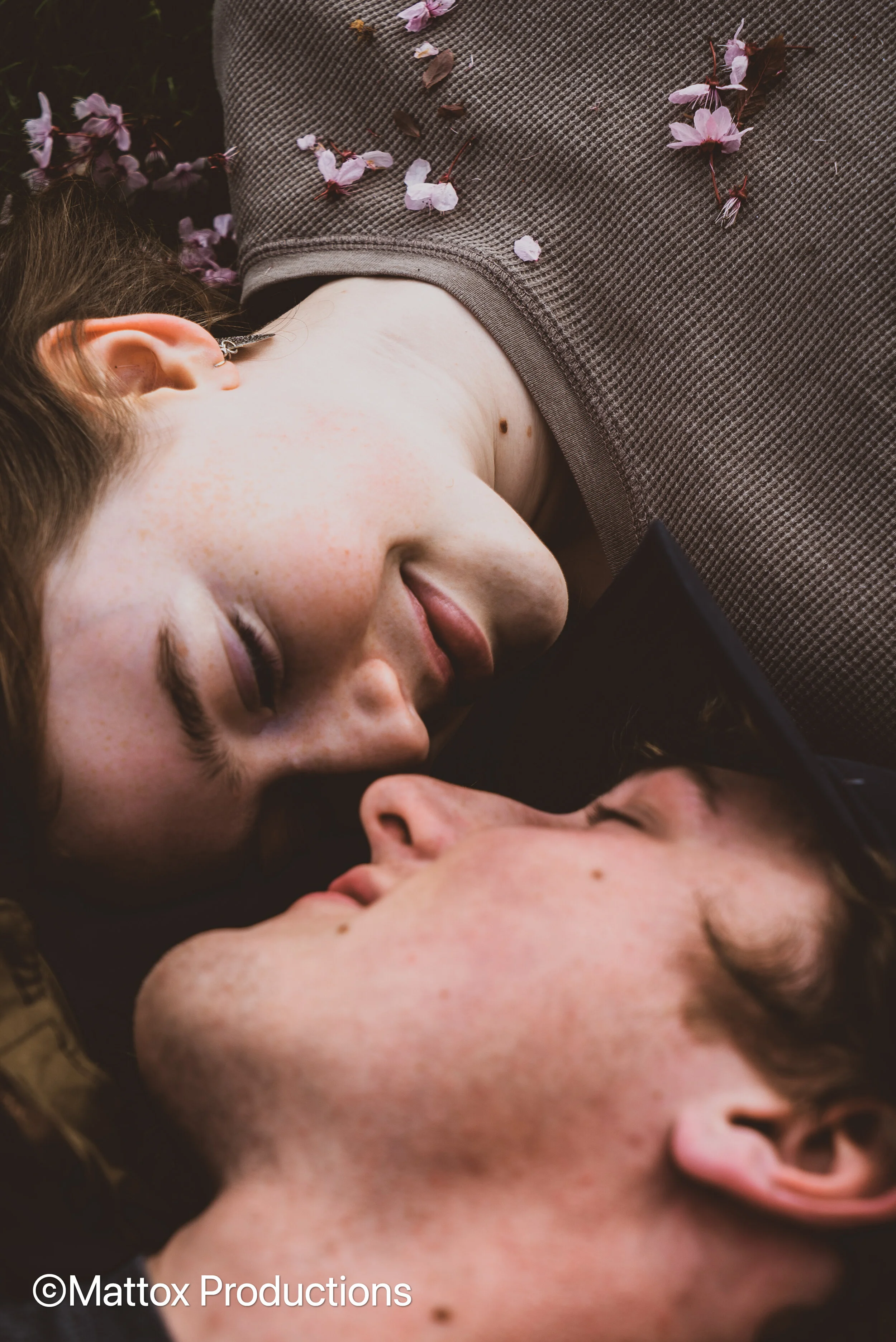 Two young people lying close together with their eyes closed, facing each other, surrounded by pink and purple blossoms on their clothing and nearby ground.