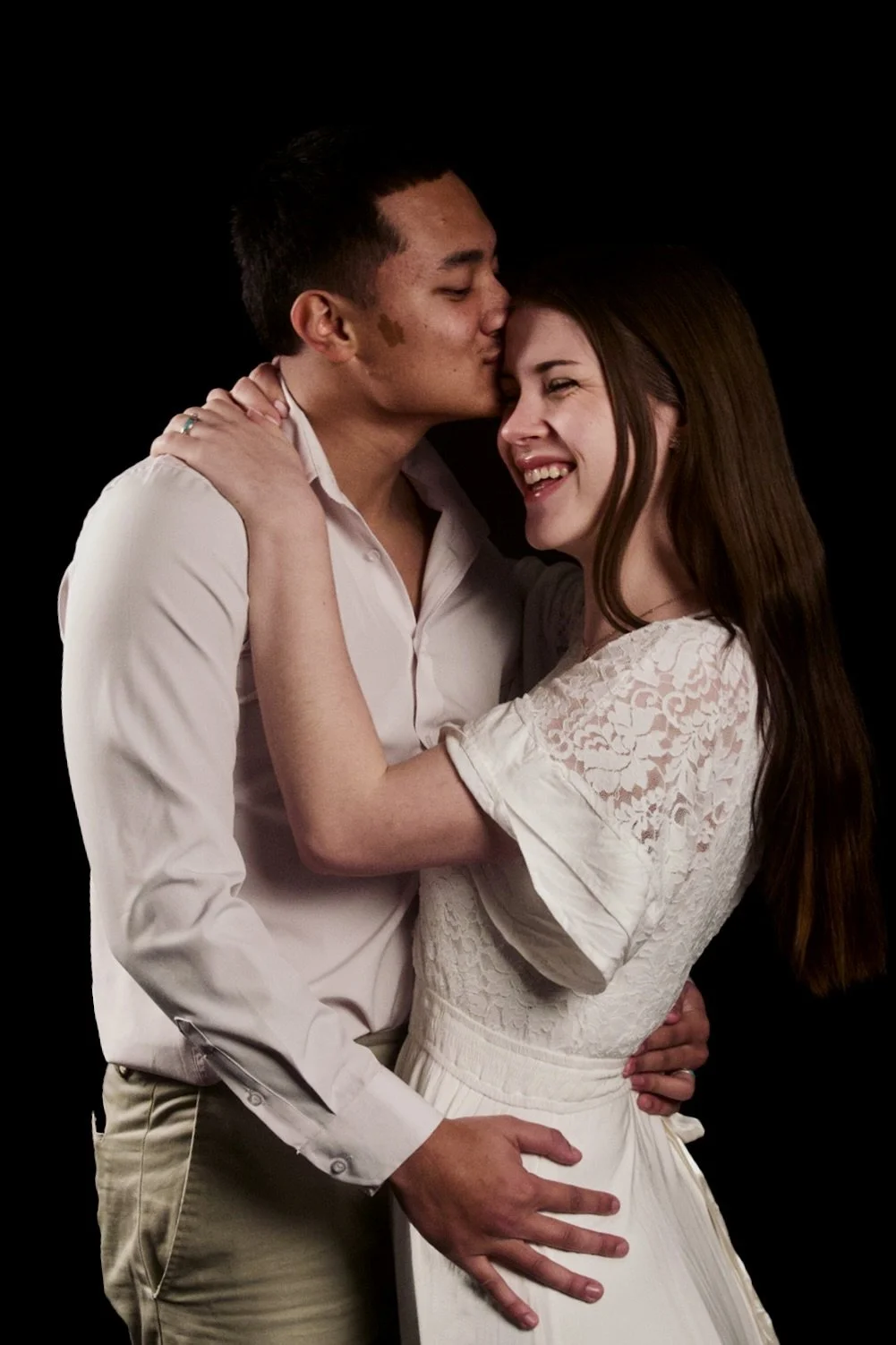 A couple embracing, with the man giving a kiss on the woman's forehead, both smiling joyfully against a black background.