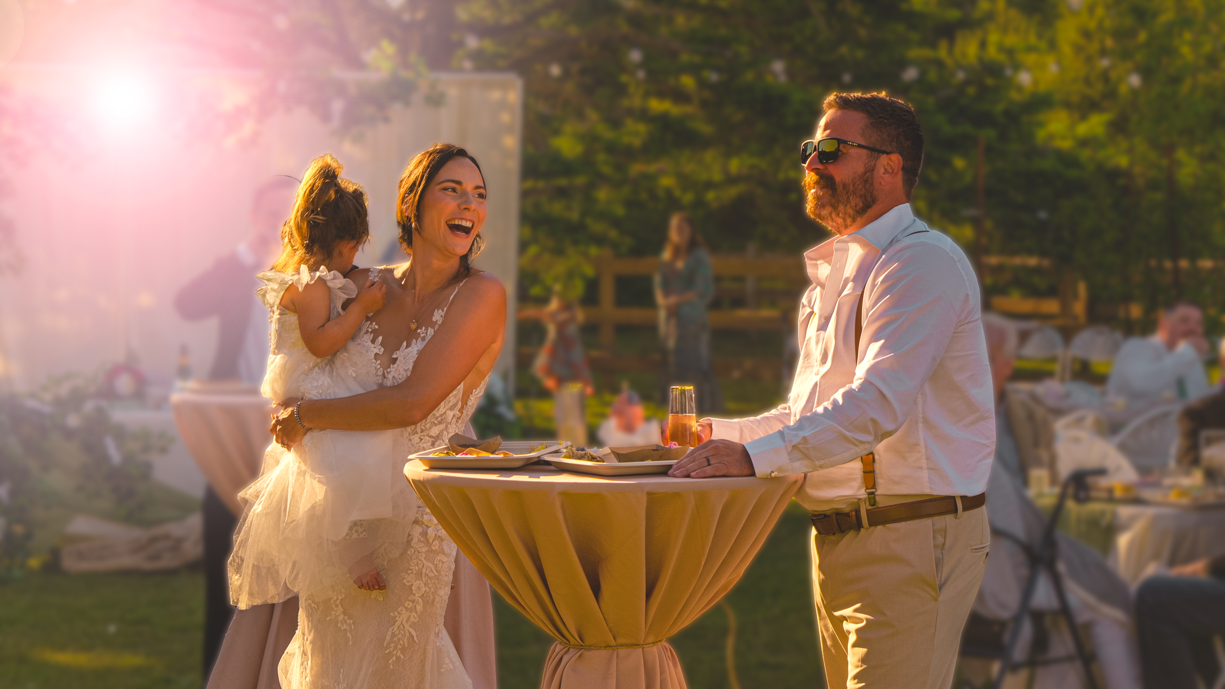 A wedding reception outdoor during sunset, with a bride holding a child, smiling, and a man in sunglasses serving drinks at a high-top table.